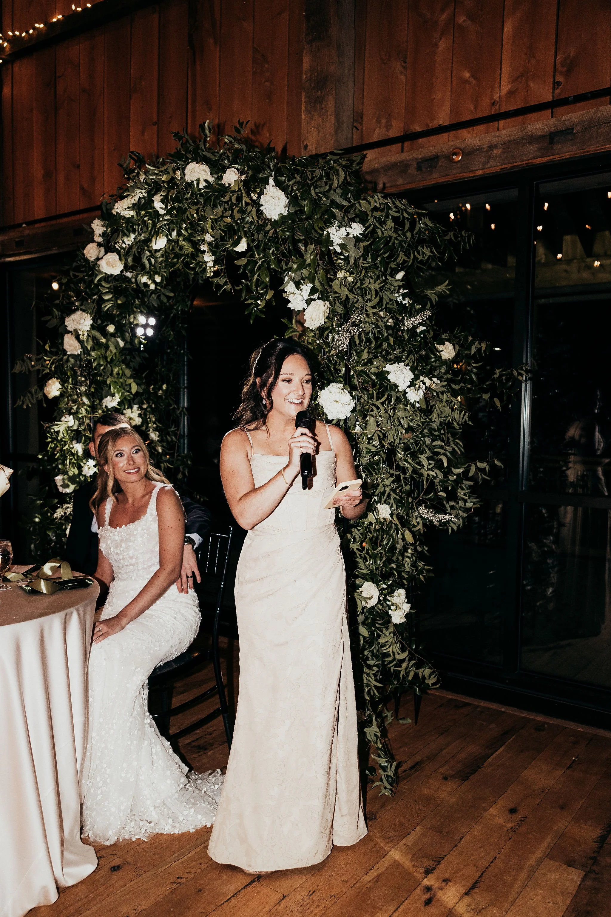 A woman giving a speech at a wedding reception, standing under a floral arch, dressed in a white wedding gown. A seated bride in a similar white gown sits to her left, and guests are visible in the background.