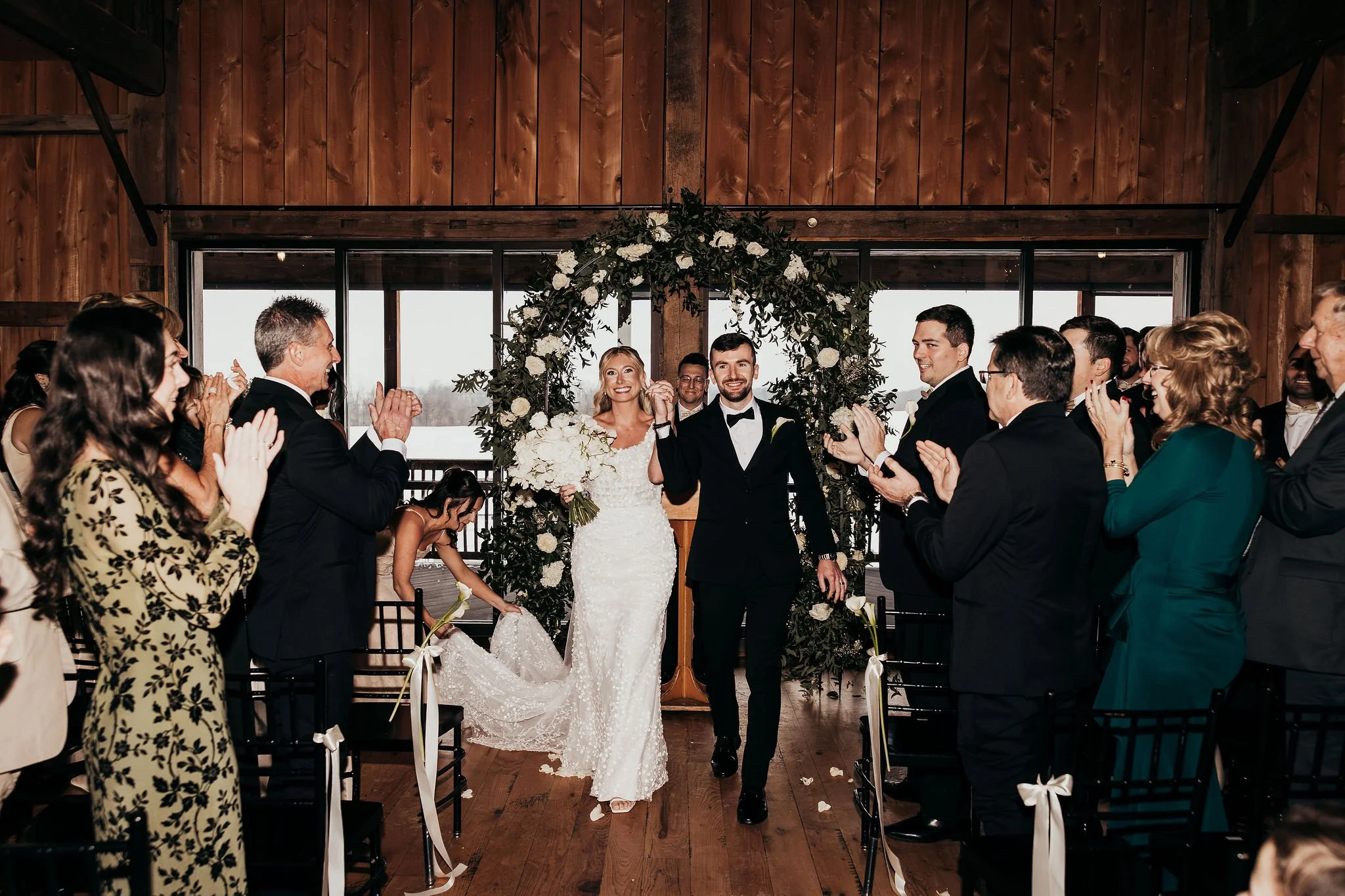 A bride and groom walking down the aisle at their wedding ceremony, surrounded by guests clapping and smiling inside a wooden venue with large windows and floral decorations.