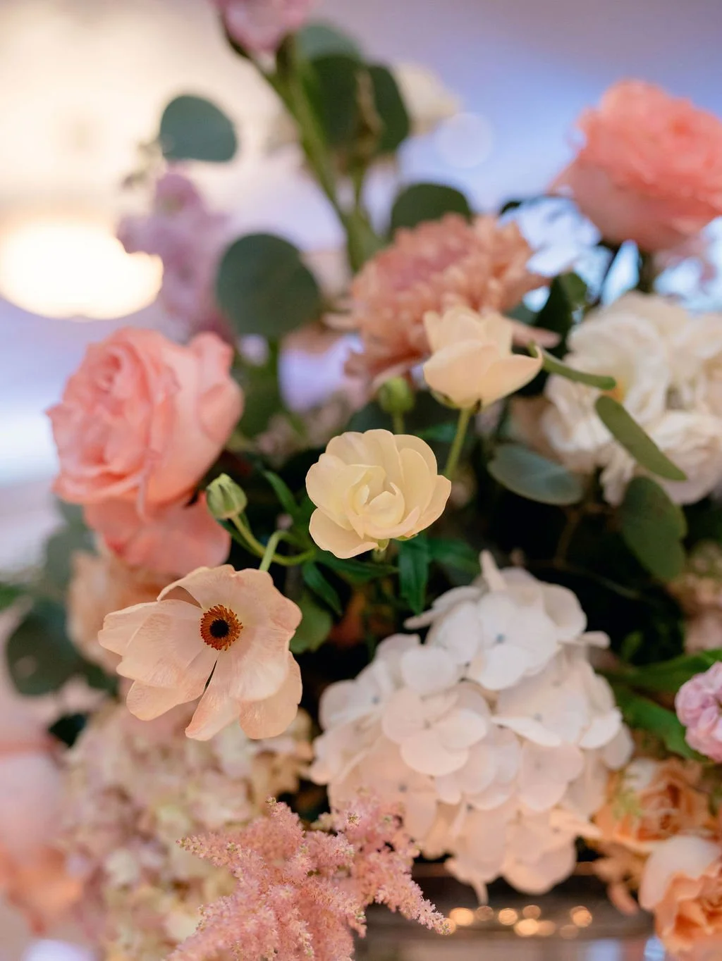 Close-up of various light pink, peach, and white flowers including hydrangeas and roses, with green leaves, in soft indoor lighting.