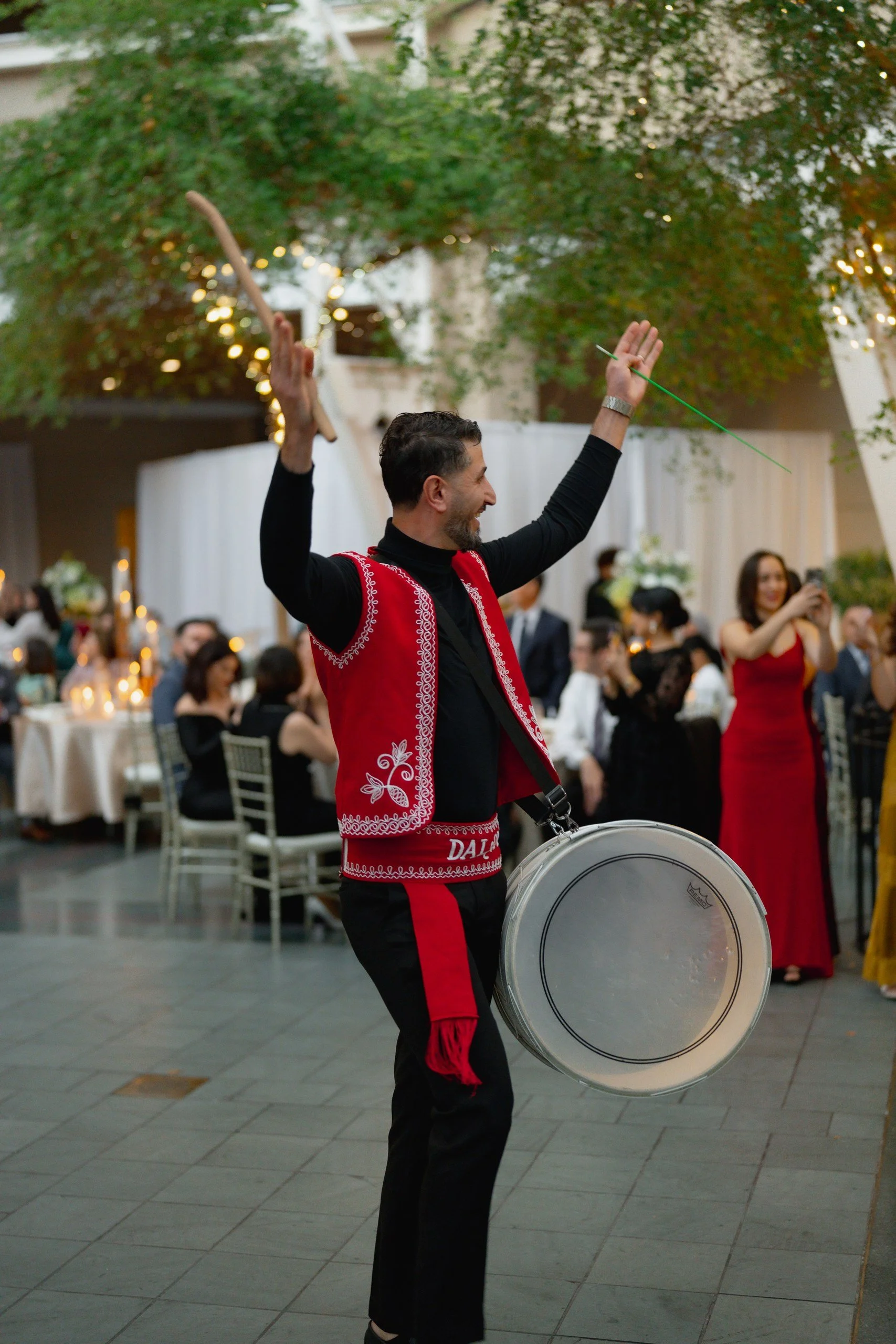 A man smiling and playing a bass drum at an indoor event, dressed in black with a red embroidered vest, surrounded by people seated at tables and standing, with festive lights and greenery in the background.