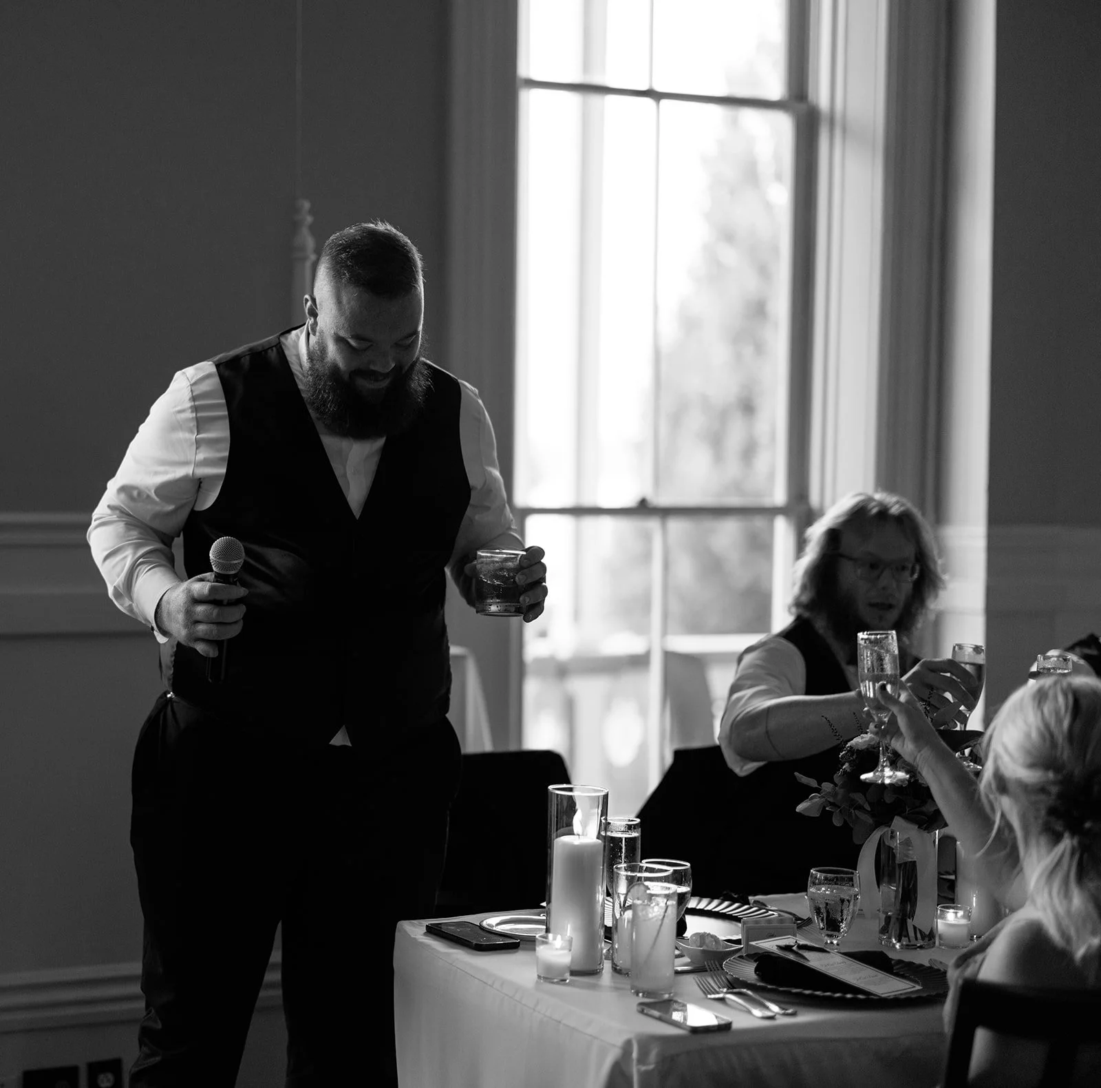 A black and white photo of a man with a beard and a buzz cut hairstyle, wearing a white shirt and a vest, holding a drink and a microphone. He is standing next to a table with candles and drinks, with wedding guests sitting and talking in the backgro
