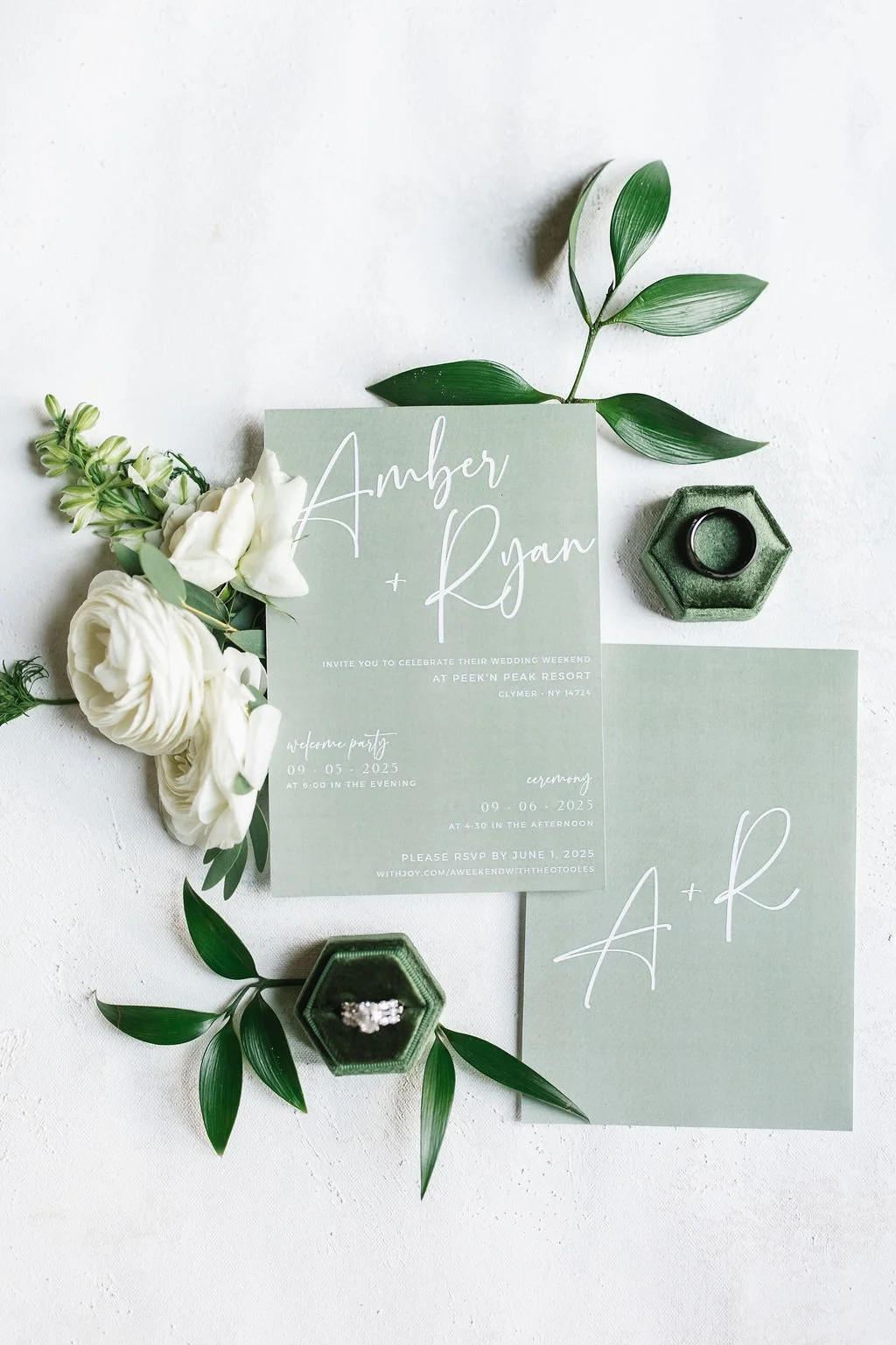 Wedding invitation cards for Amber and Ryan, decorated with white flowers and green leaves, with rings in a green hexagonal box.