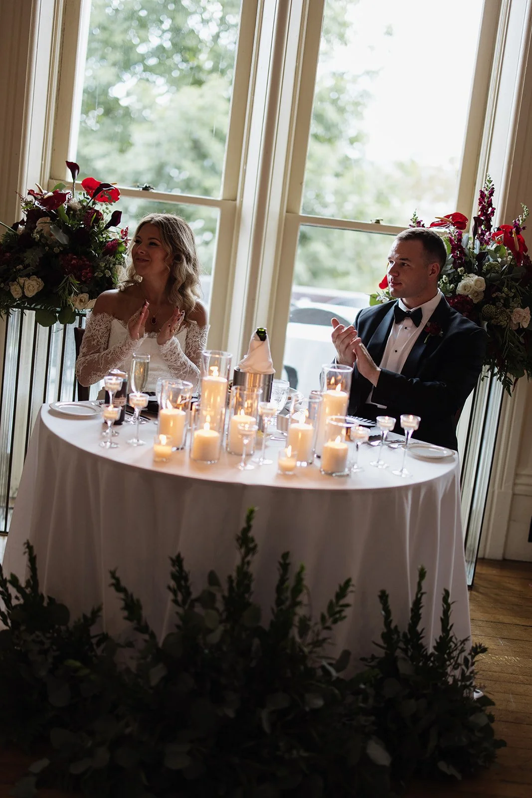 A newlywed couple sitting at a decorated table with lit candles, surrounded by floral arrangements, during their wedding reception.