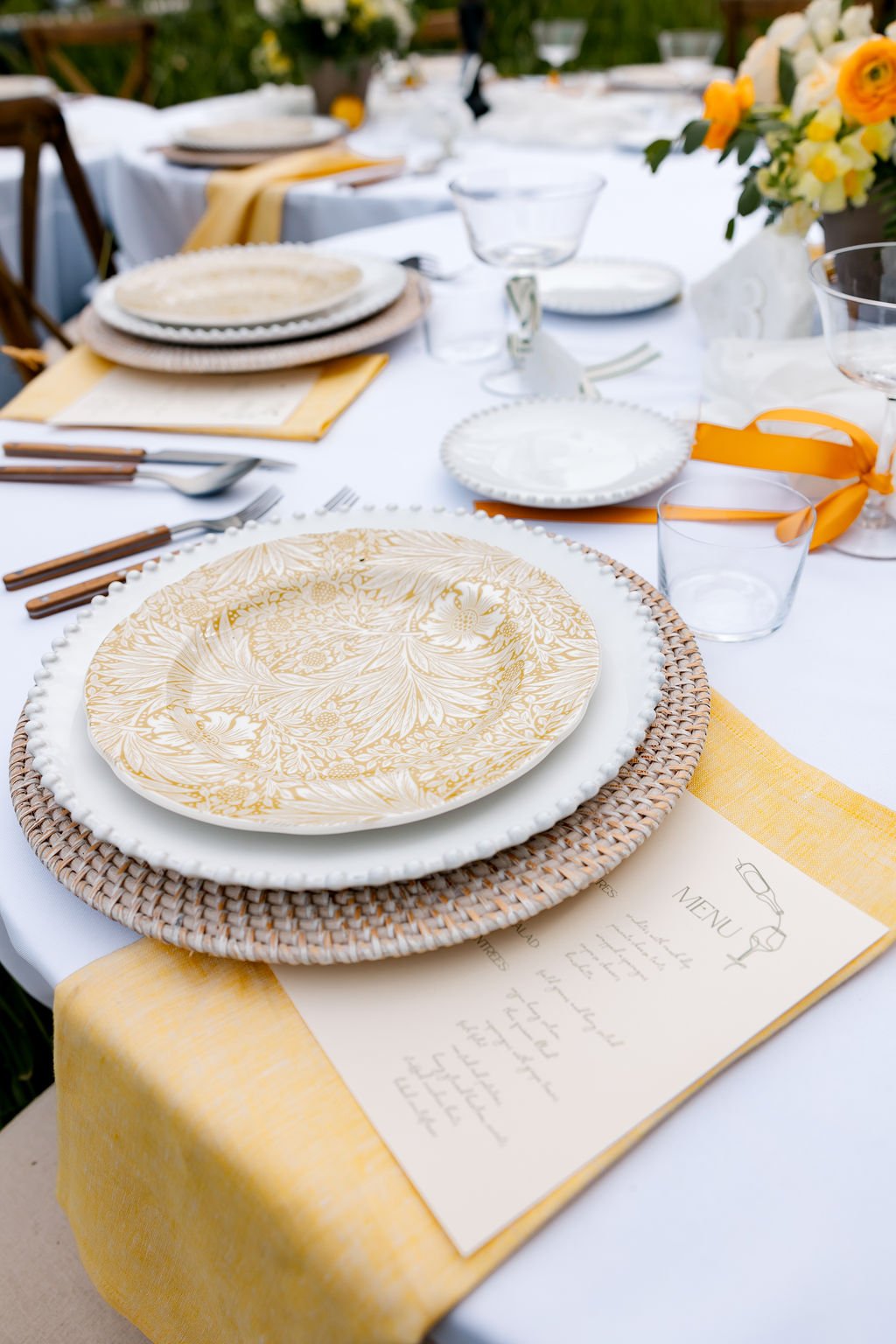 Elegant outdoor table setup with white and gold patterned plates, woven chargers, glassware, utensils, a menu, and yellow flowers.