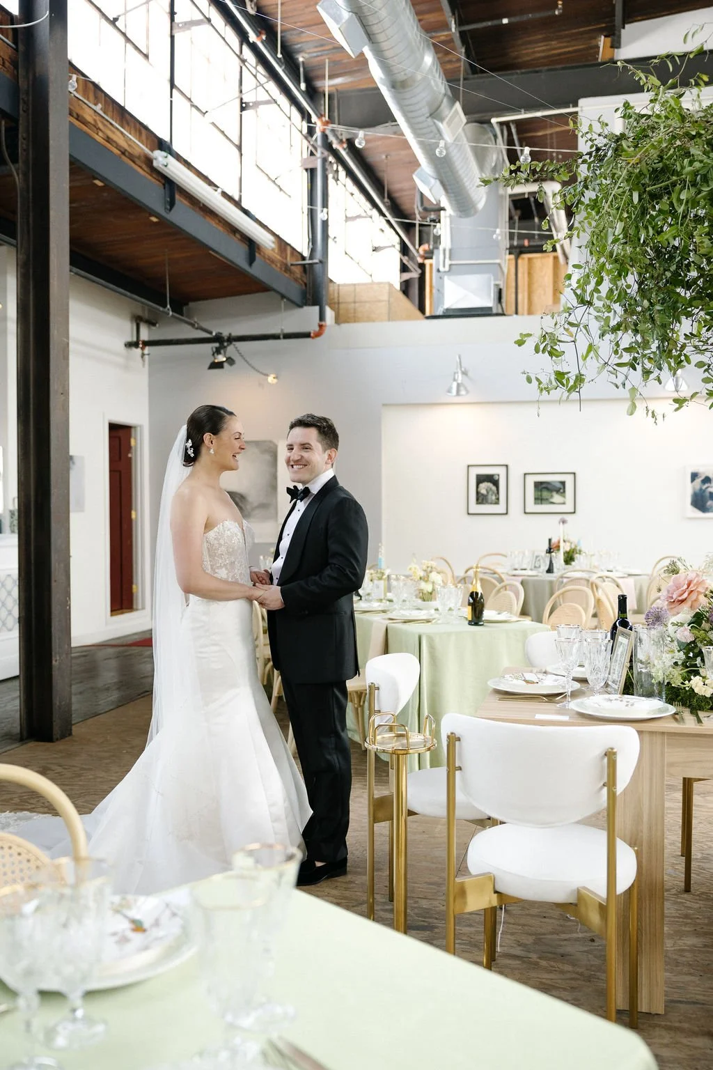 A bride and groom holding hands and smiling at each other inside a decorated wedding reception space with tables set for a celebration.