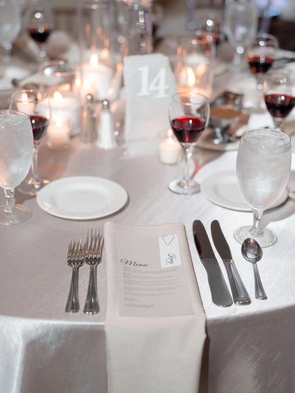 Formal dining table setup with white tablecloth, wine glasses filled with red wine, water glasses, plates, silverware, a menu, a centerpiece with candles, and a table number 14 sign.
