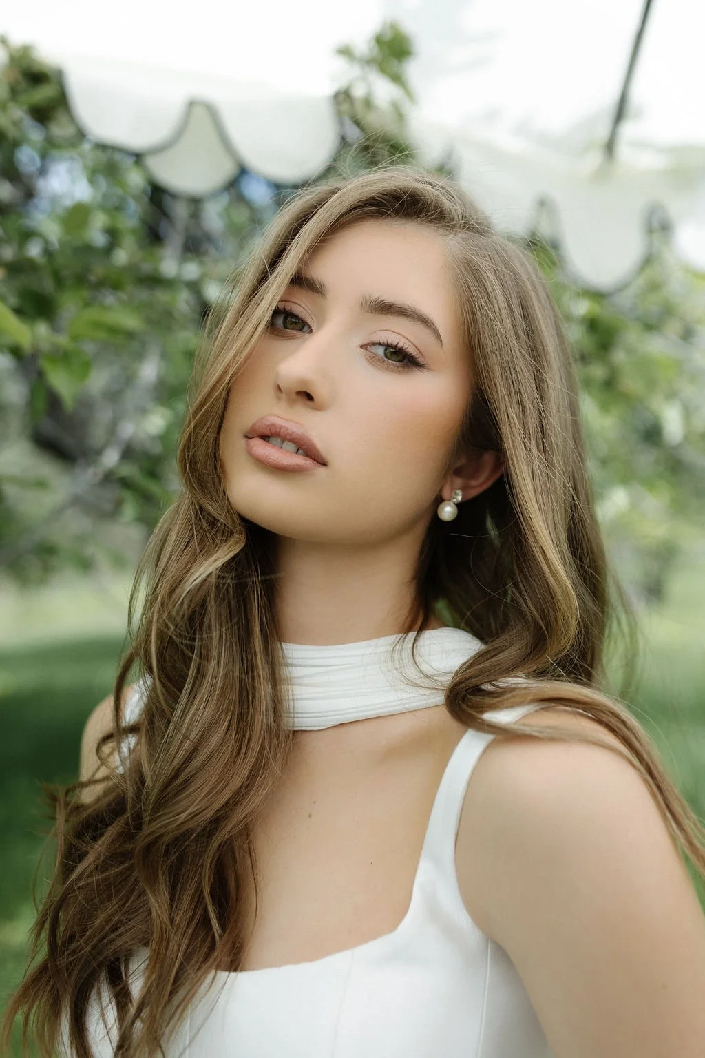 A young woman with long wavy light brown hair and light skin, wearing pearl earrings and a white sleeveless top with a high collar, standing outdoors with greenery in the background.