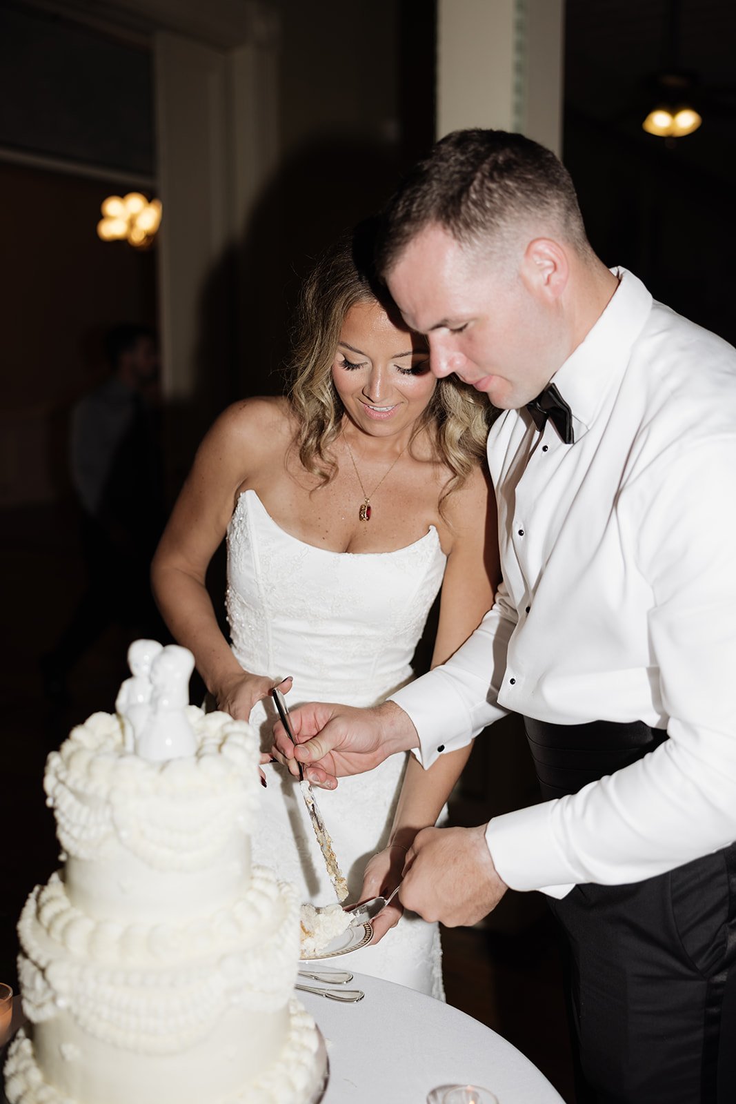 A bride and groom cutting a wedding cake together at their wedding reception, with the bride wearing a white strapless dress and the groom in a white shirt and black bow tie.