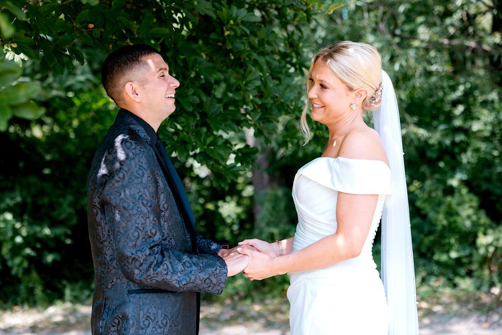 A bride and groom holding hands and smiling at each other outdoors with green foliage in the background.