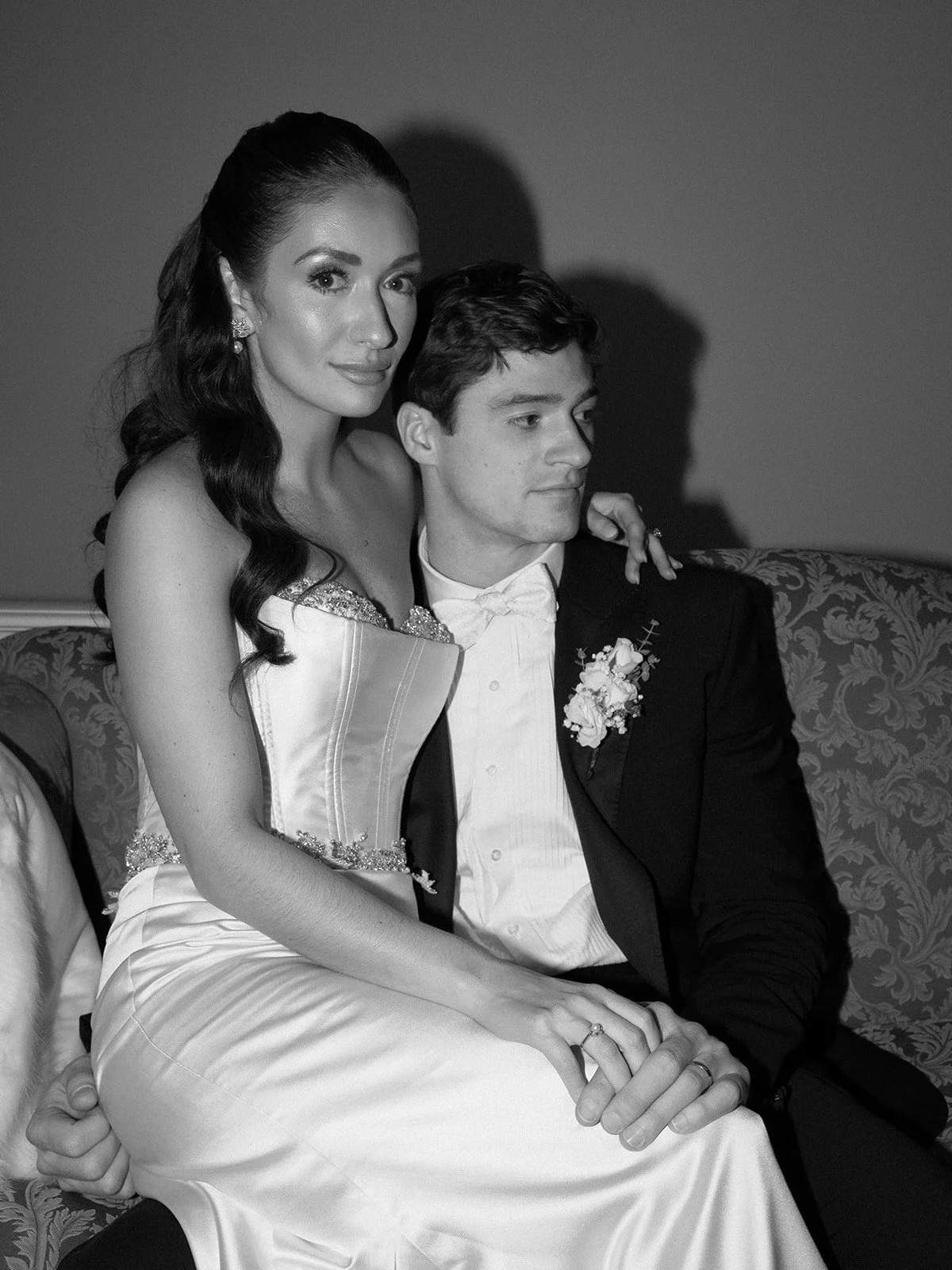 A black and white photo of a woman in a wedding dress and a man in a tuxedo sitting close together on an ornate sofa. The woman has dark, wavy hair and is wearing earrings, while the man has short hair and is wearing a boutonnière.