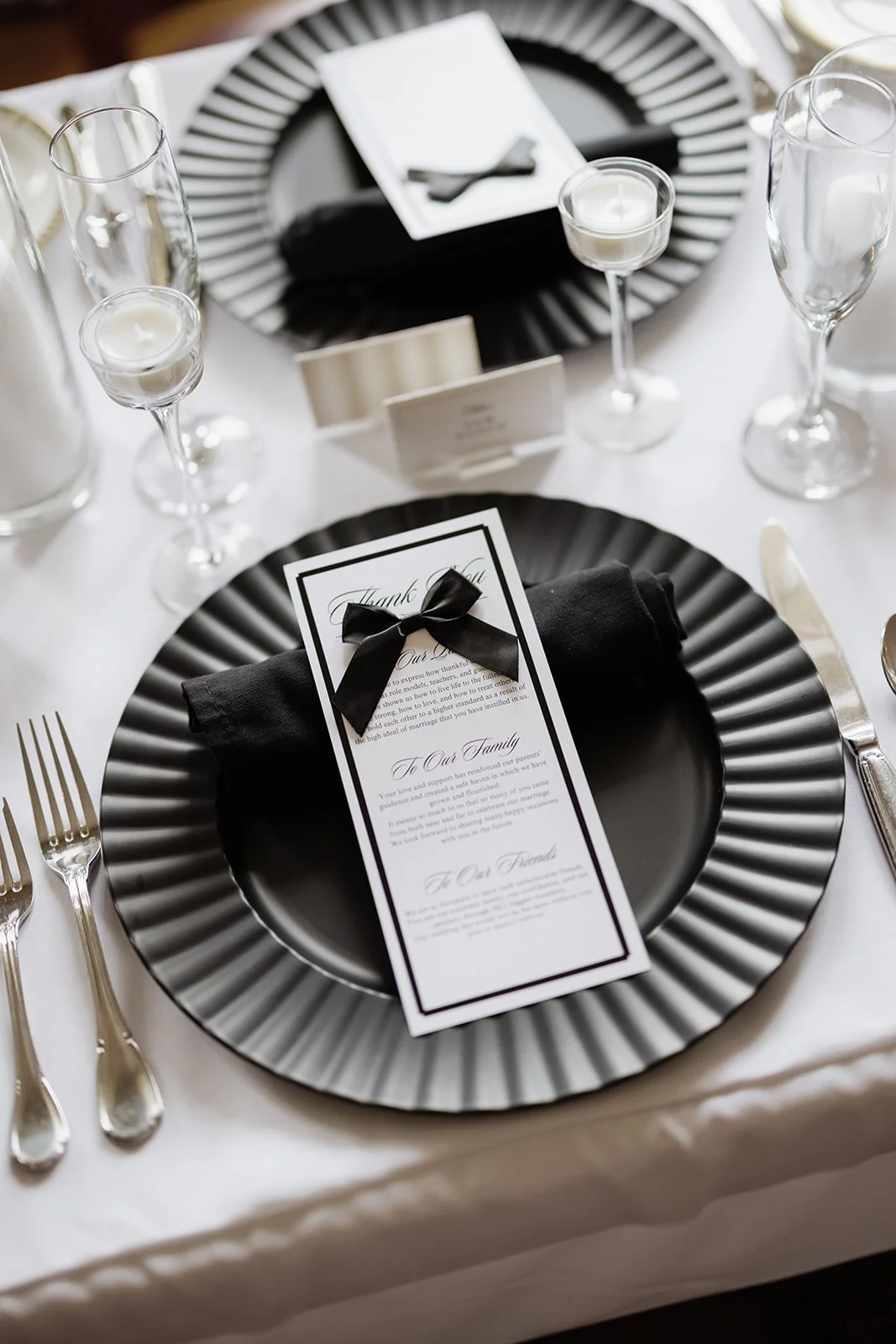 Elegant table setting with black and white color scheme, featuring a black plate with a black napkin and a thank you card tied with a black ribbon, surrounded by wine glasses, candles, and cutlery.