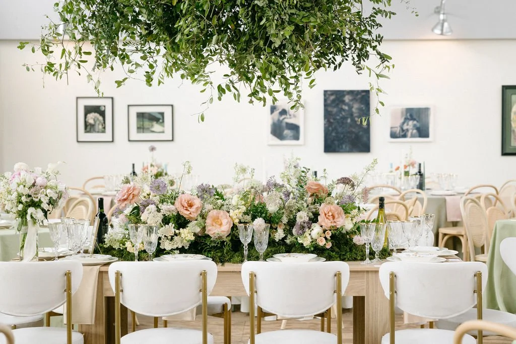 Elegant dining setup with floral centerpieces, glassware, and chairs, decorated for a celebration or event, with greenery hanging from the ceiling.