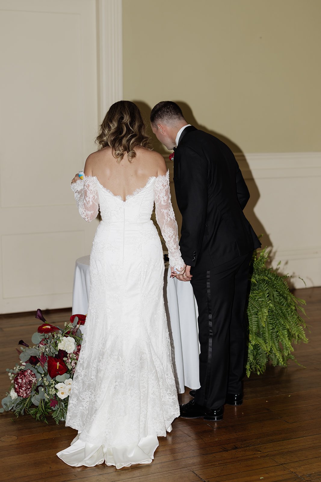 A bride and groom holding hands during their wedding ceremony, standing near a floral arrangement on a wooden floor.