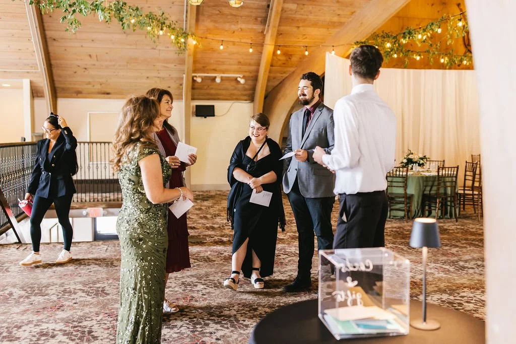Group of five adults standing and talking in a decorated event space with wooden ceiling beams, string lights, and green tablecloths.