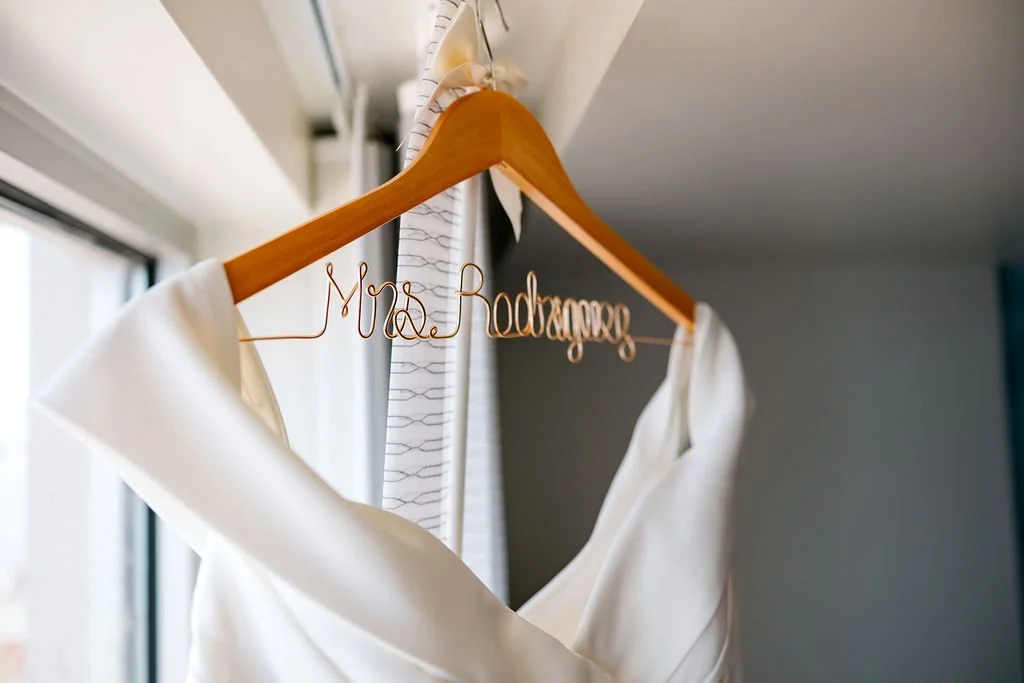 White wedding dress hanging on a wooden hanger inside a room with natural light, with a custom wire hanger that spells out 'Mrs. Rodriguez'.