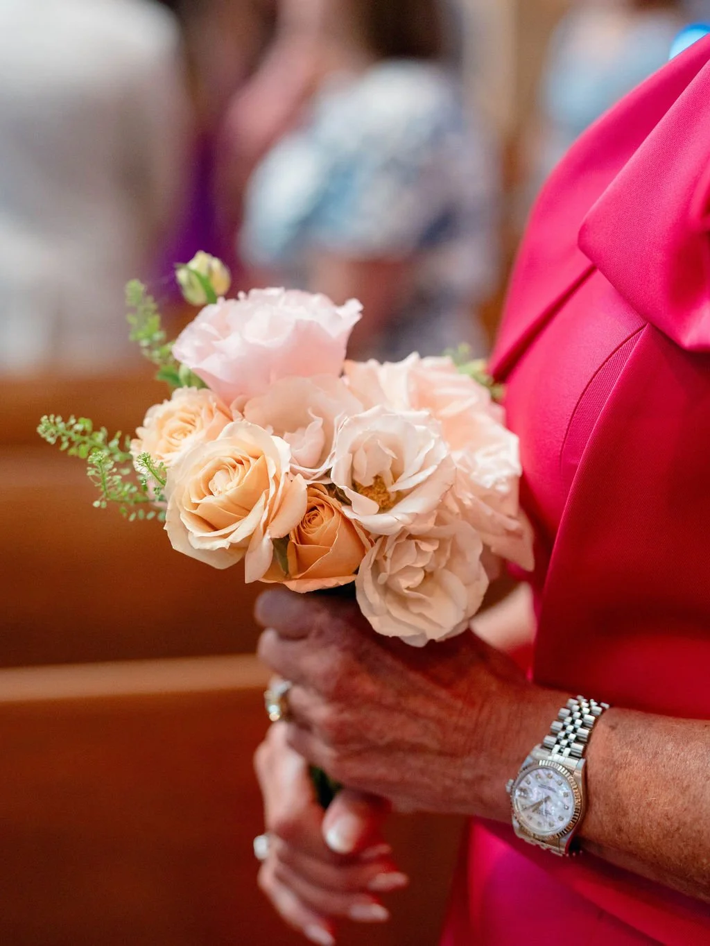 A woman in a bright pink dress holding a bouquet of light pink and peach roses with some green foliage, wearing a silver watch and a ring.