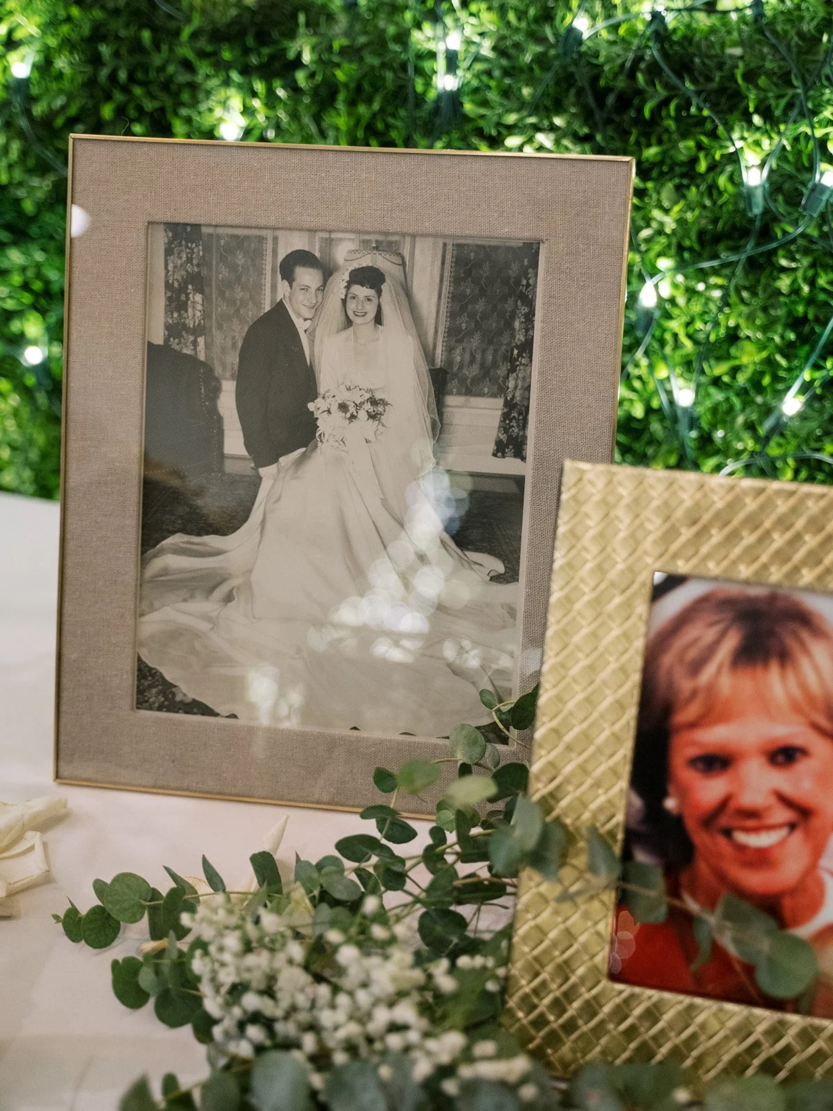 A black-and-white wedding photo of a bride and groom in a fancy room with floral curtains, displayed in a beige frame, surrounded by green leaves and small white flowers. A small photo of a woman with short blonde hair and a red outfit is in a gold w