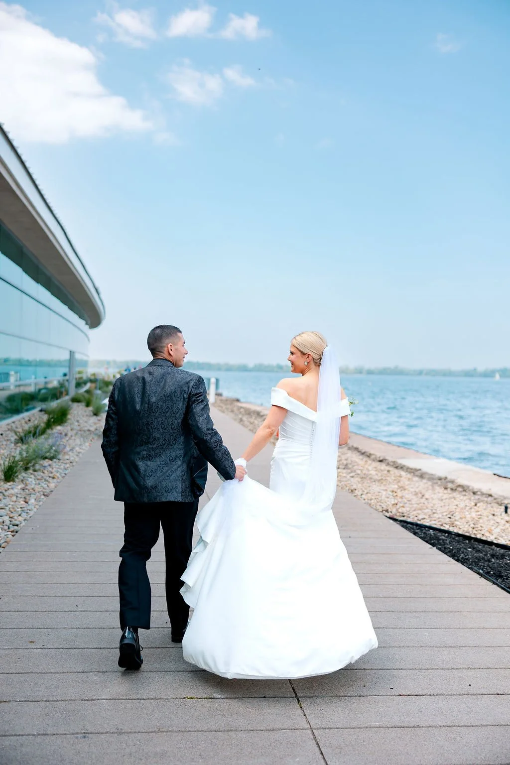A bride and groom holding hands walking along a waterfront promenade on a sunny day.