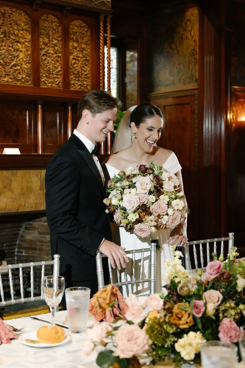 A bride and groom standing together at their wedding reception, smiling and holding a large bouquet of flowers, surrounded by floral arrangements and table settings.
