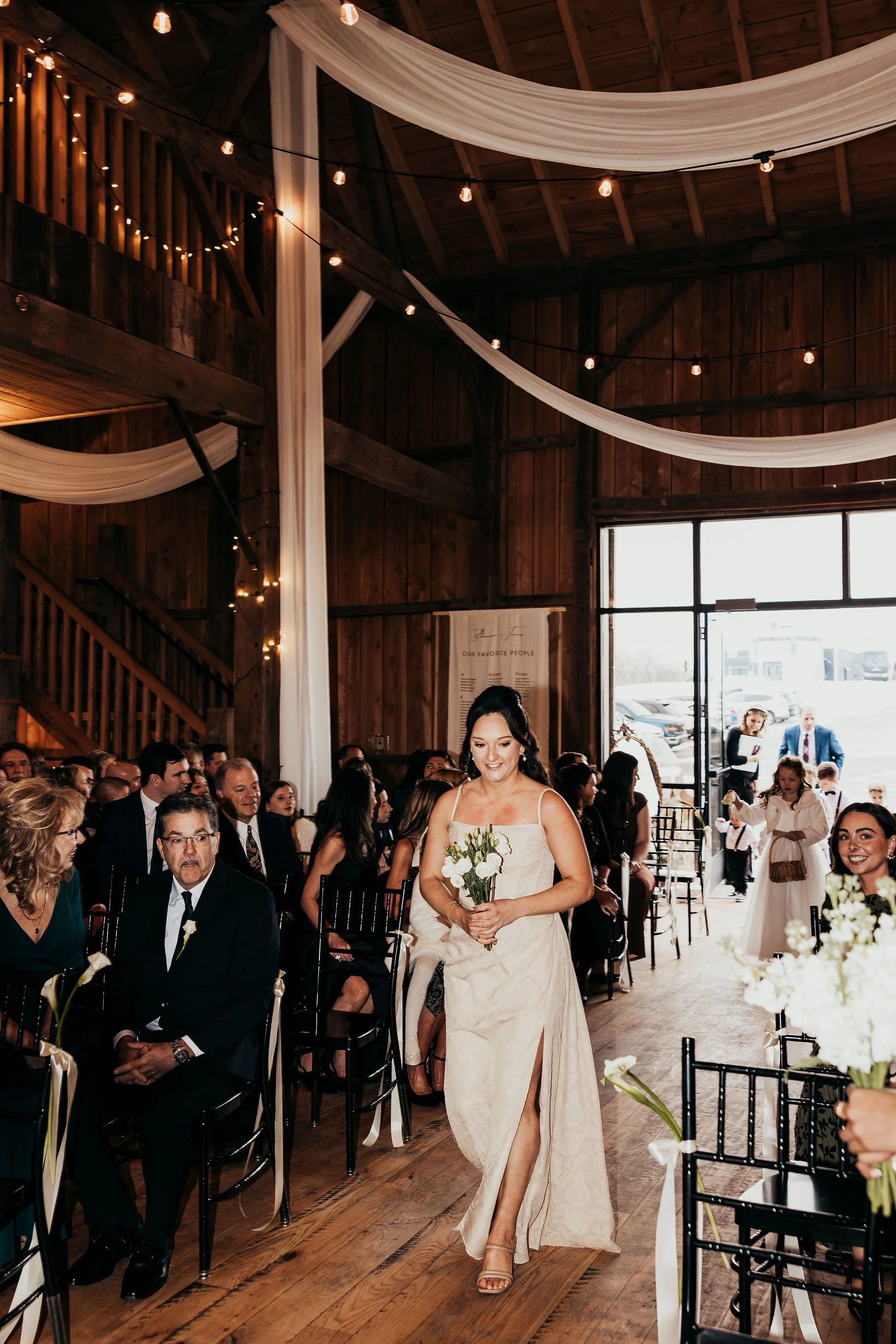 A woman in a beige wedding dress holding a bouquet walks down the aisle during a wedding ceremony inside a wooden barn decorated with draped fabric and string lights. Guests seated on either side are watching her.