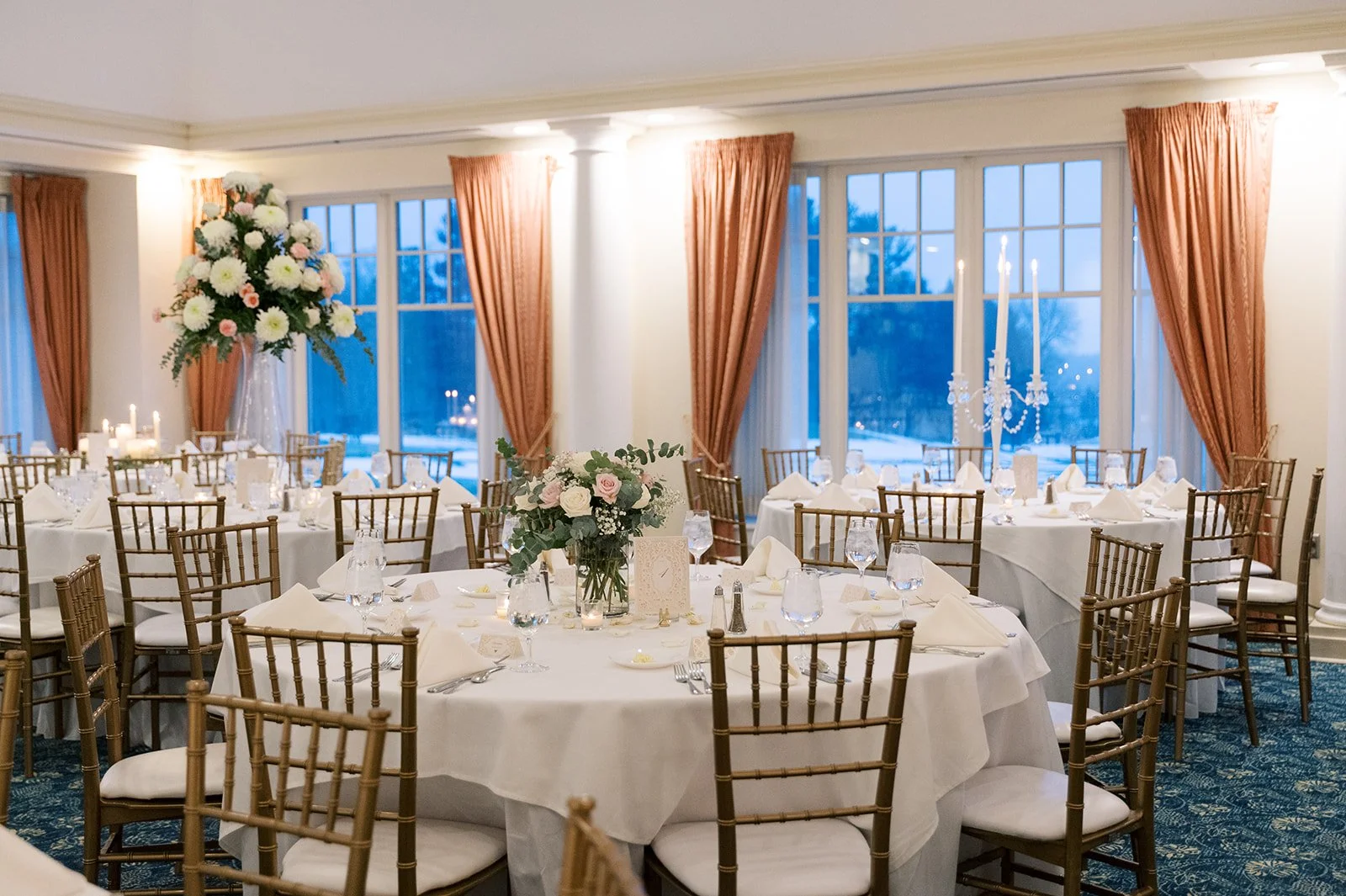 Elegant banquet room with round tables draped in white tablecloths, adorned with floral centerpieces, candlelit candelabras, and gold chairs, large windows with orange curtains overlooking a wintery landscape.