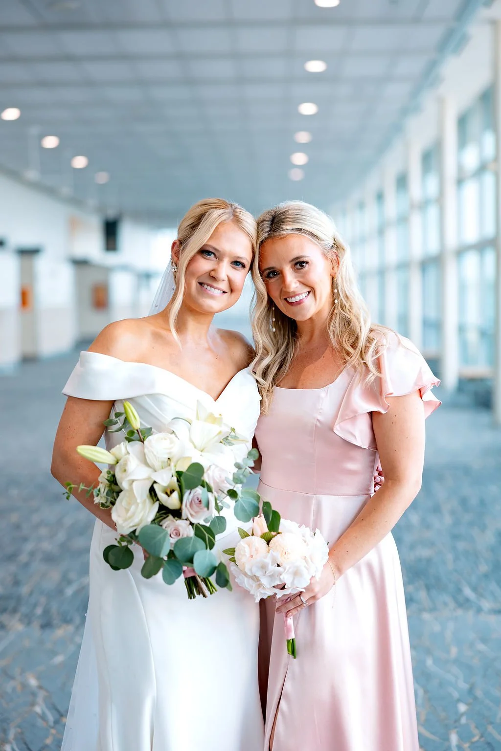 Two women, one in a wedding dress and the other in a pink dress, smiling and holding bouquets, standing in a bright indoor space with large windows.
