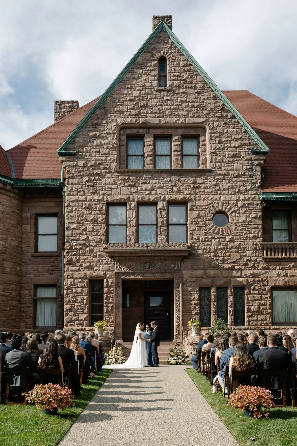 Wedding ceremony in front of a large stone building with a red roof, groom and bride exchanging vows under a small arch, guests seated on either side, floral arrangements on the ground.