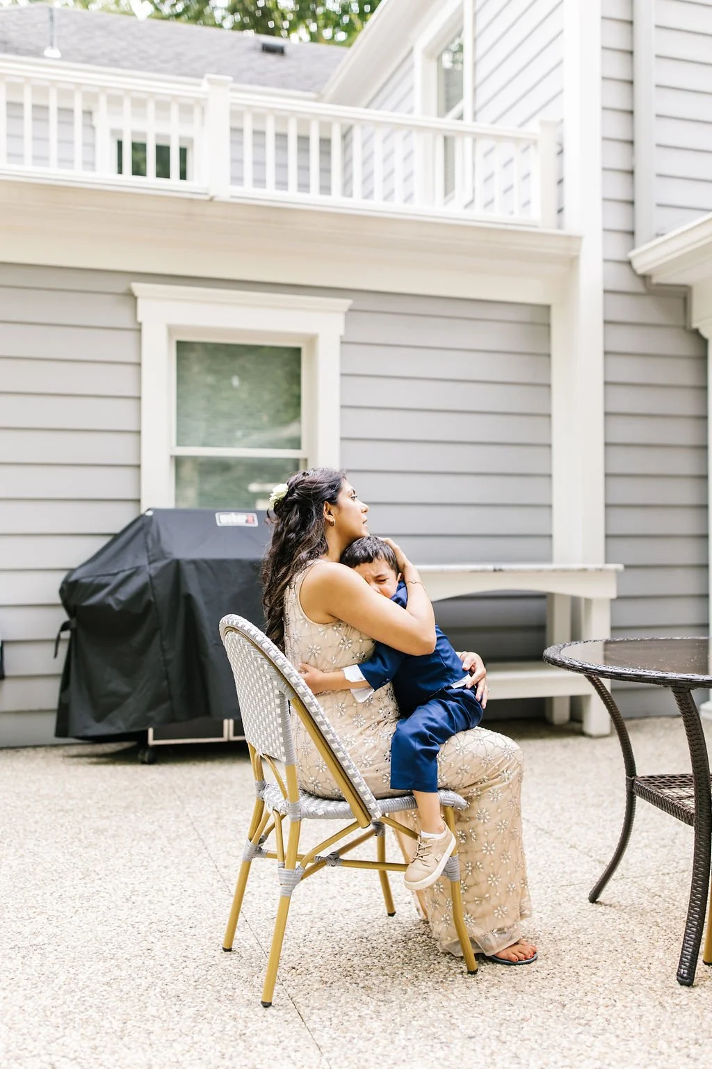 A woman and a young boy sitting outside, with the woman hugging the boy, who appears to be crying or upset, on a patio area near a house with grey siding.