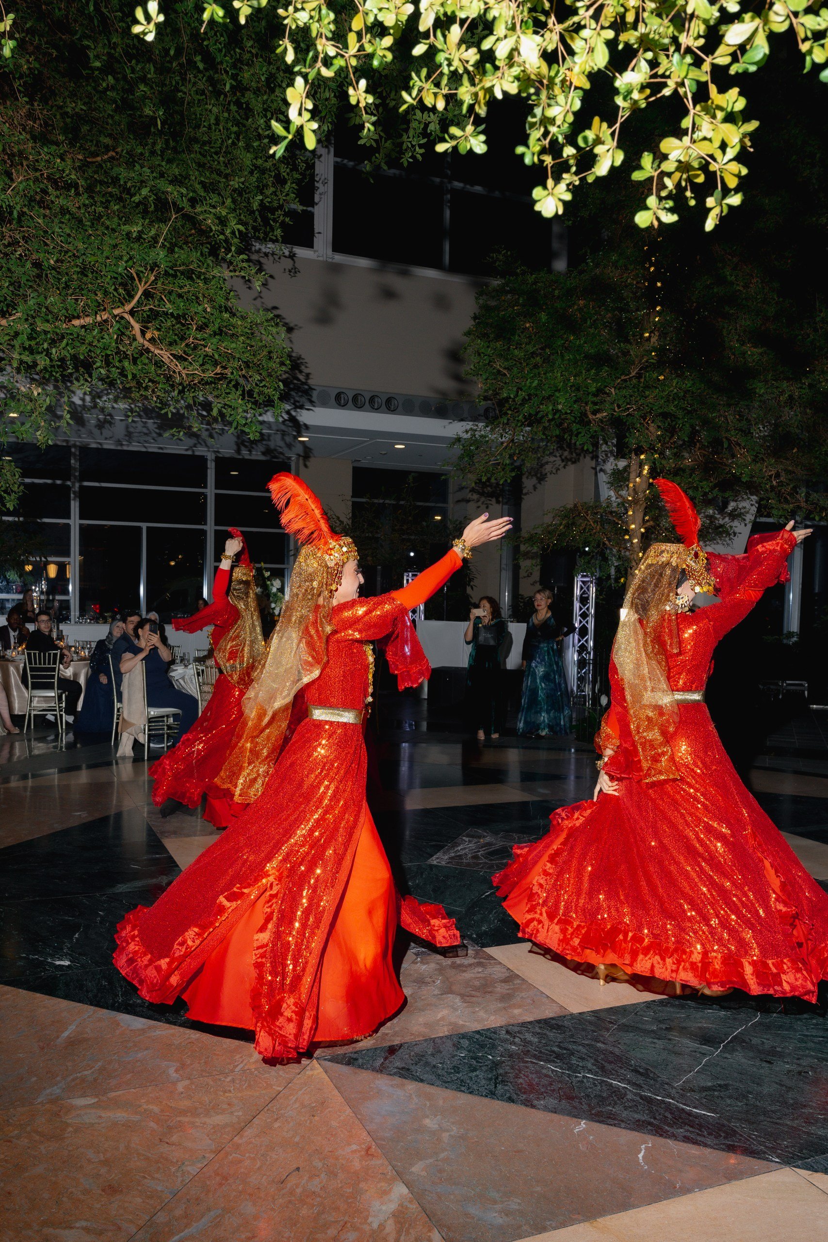 Three women dressed in vibrant red and gold costumes with headpieces and flowing skirts are performing a traditional dance indoors. Audience members are seated at tables, watching the performance.