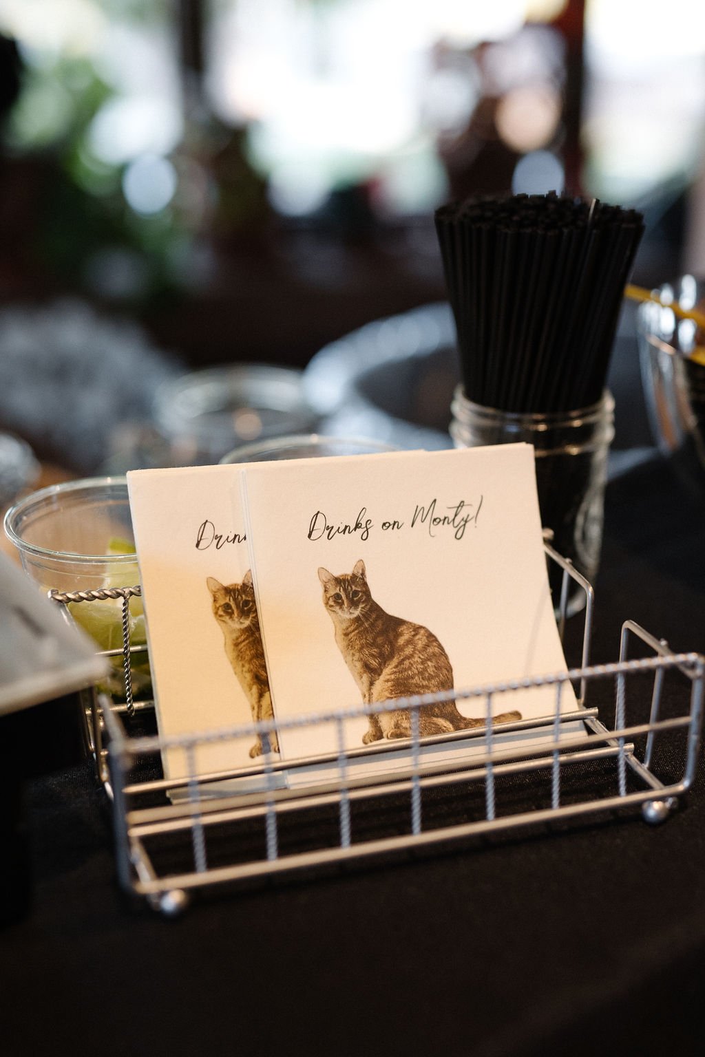 A wire basket holding paper coasters with a picture of a cat and the caption "Drinks on Monty!" in a restaurant or bar. There is a jar with black straws behind the basket.