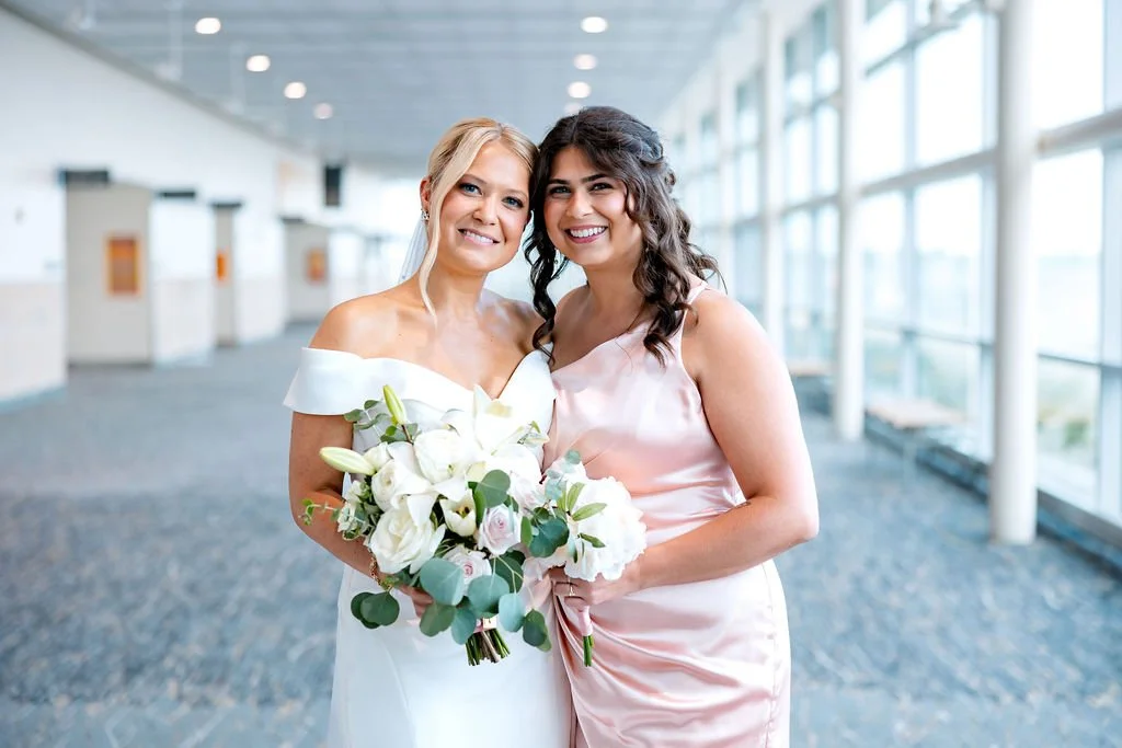 Two women in an indoor hallway, one in a wedding dress holding a bouquet of white flowers, and the other in a pink dress. They are smiling and standing close together.