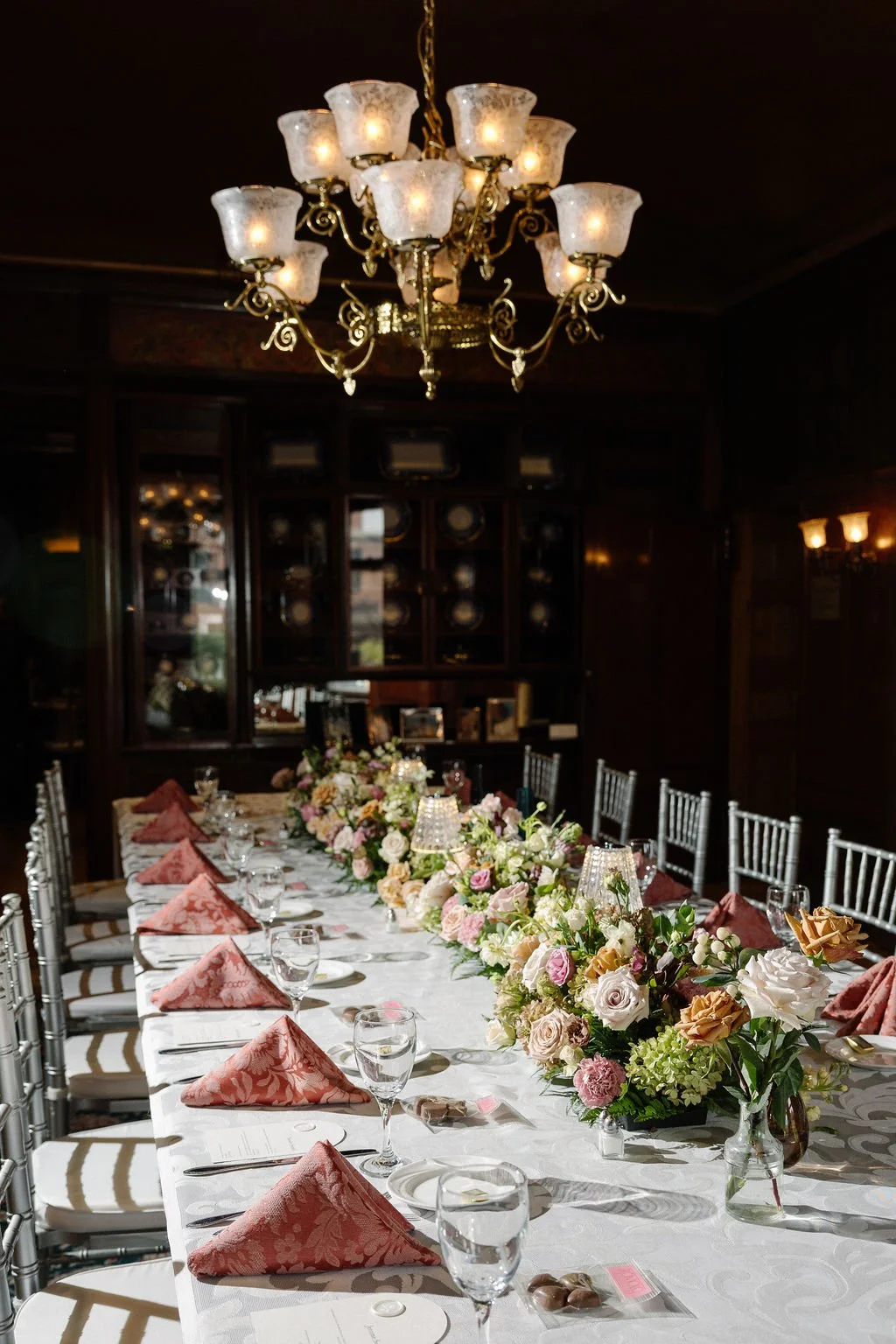 Elegant dining table set with pink napkins, floral centerpieces, glassware, and silverware, illuminated by a chandelier in a dark wood-paneled room.
