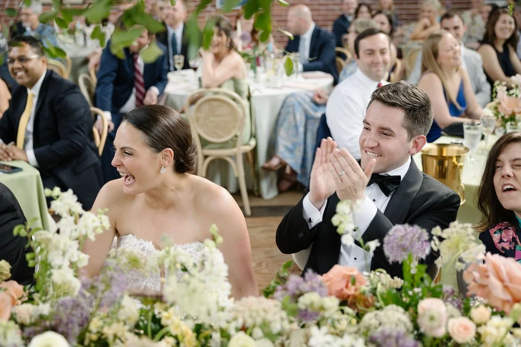 A joyful bride and groom seated at a wedding reception, surrounded by guests, with flowers and table settings visible.