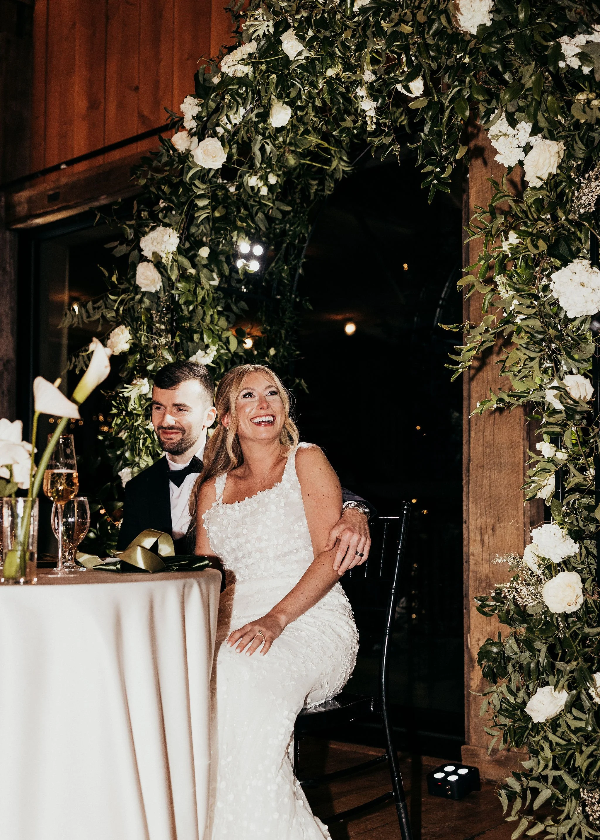 A bride and groom sitting together at their wedding reception, with the bride wearing a white wedding dress and the groom in a black tuxedo, surrounded by floral decorations and greenery.