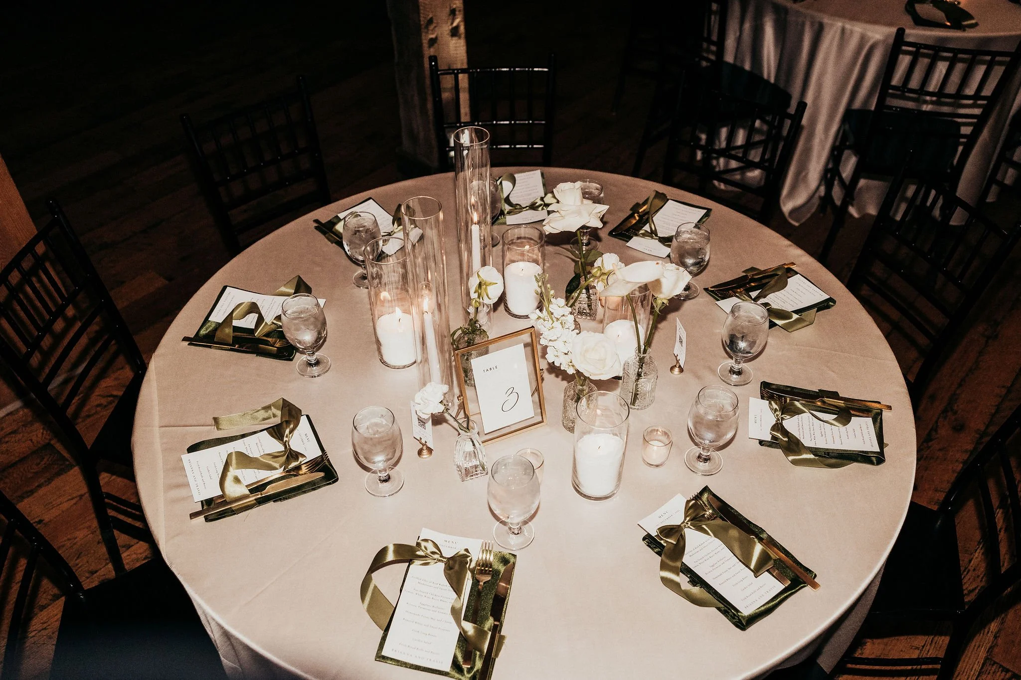 Round banquet table decorated with white flowers, candles, glassware, and gold-colored utensils, set for a formal event.