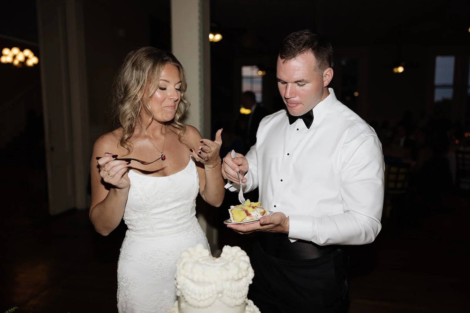 A newlywed couple, the bride in a white dress and the groom in a tuxedo with a black bowtie, sharing cake at their wedding reception.