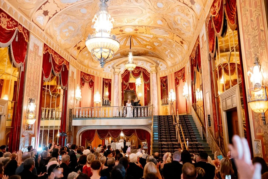 Elegant event in a grand, decorated theater with ornate ceiling and walls, a chandelier, red velvet drapery, a staircase, and a crowd of people, some taking photos.