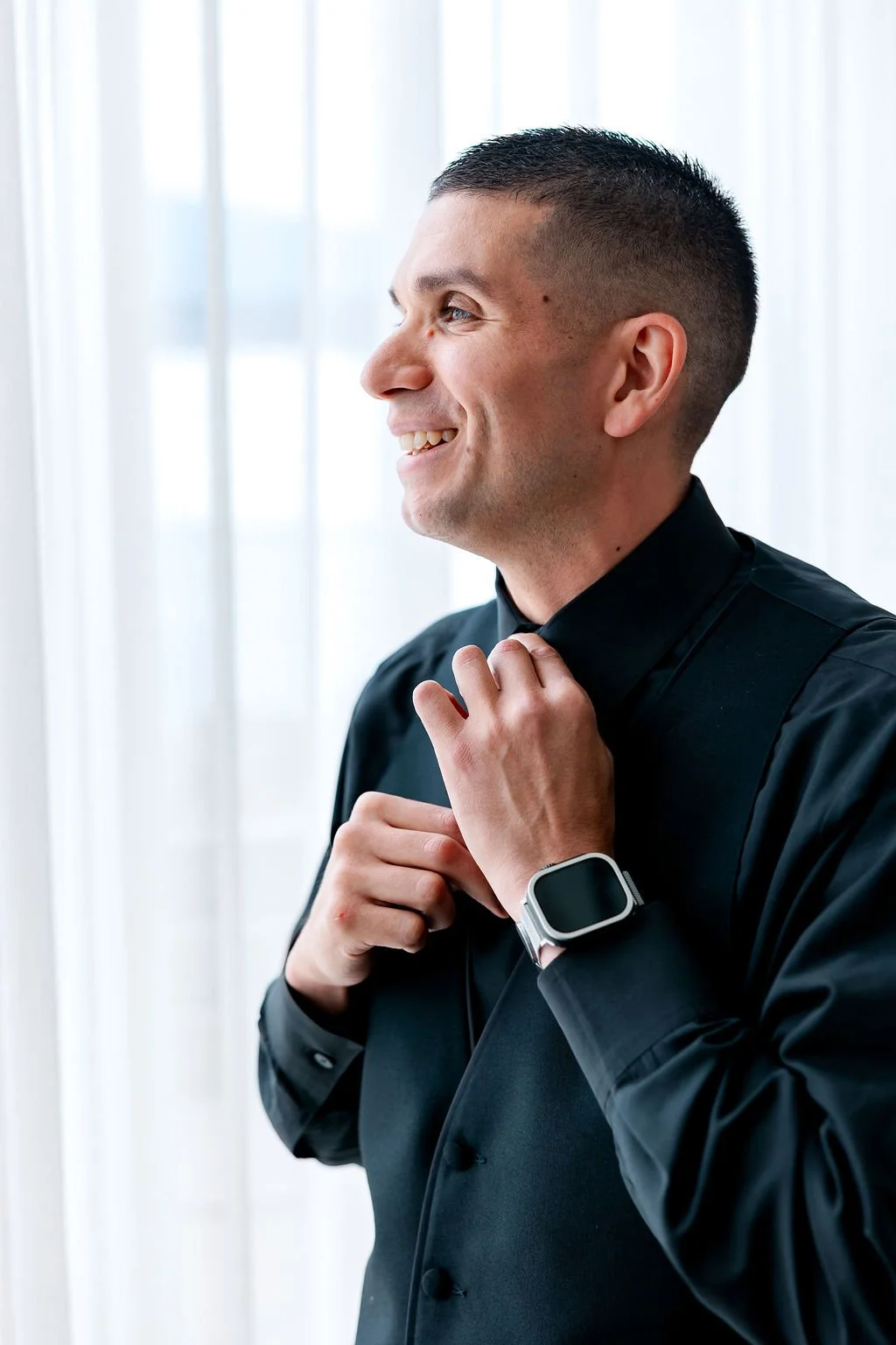 A young man adjusts his black tie while smiling, wearing a black suit and smartwatch, in front of white curtains.