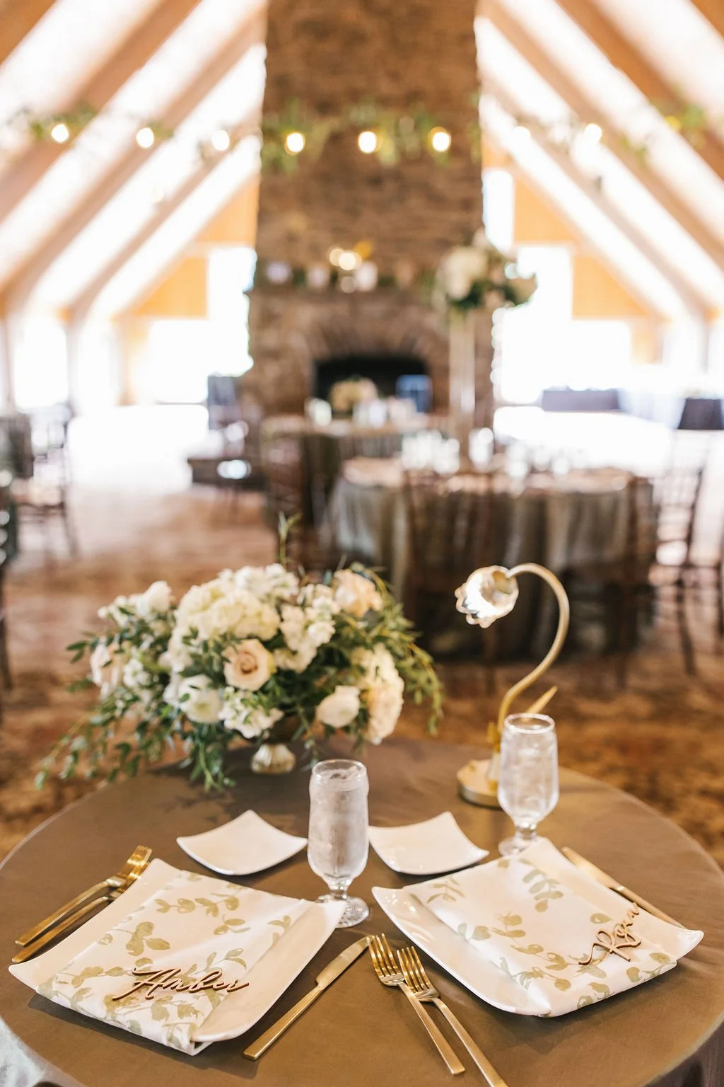 Wedding reception table with floral centerpiece, gold cutlery, napkins with a guest's name, and two glasses of water, in a rustic venue with a stone fireplace and string lights.
