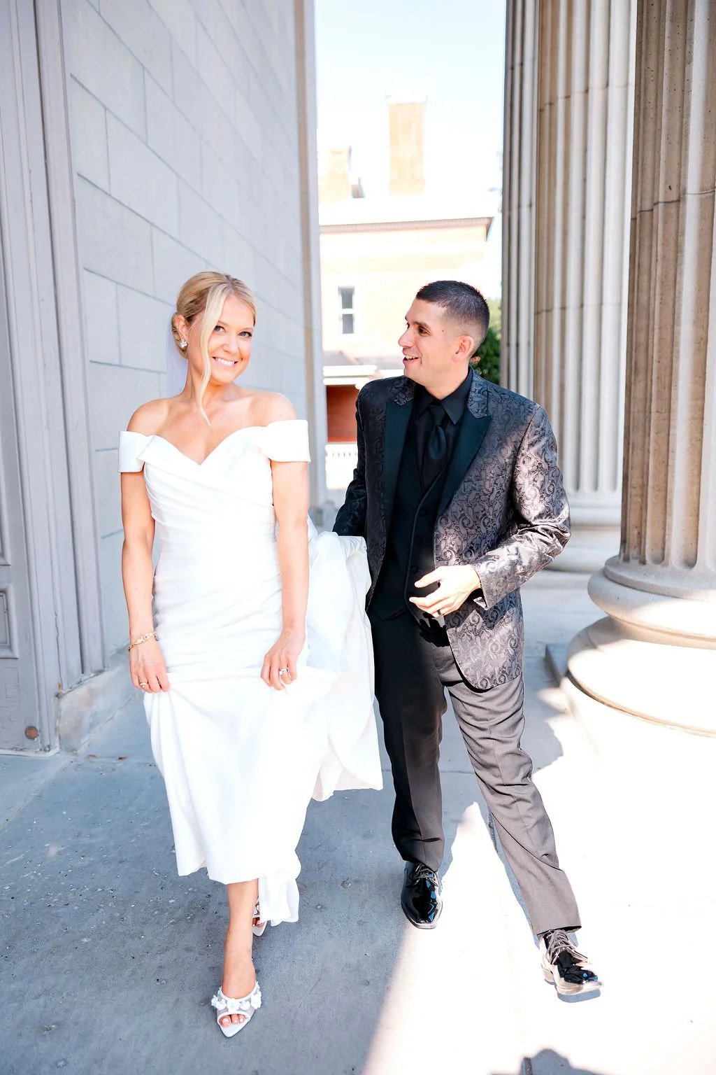 A bride and groom walking together outdoors, smiling and looking at each other, with the bride in a white wedding dress and the groom in a dark patterned suit.