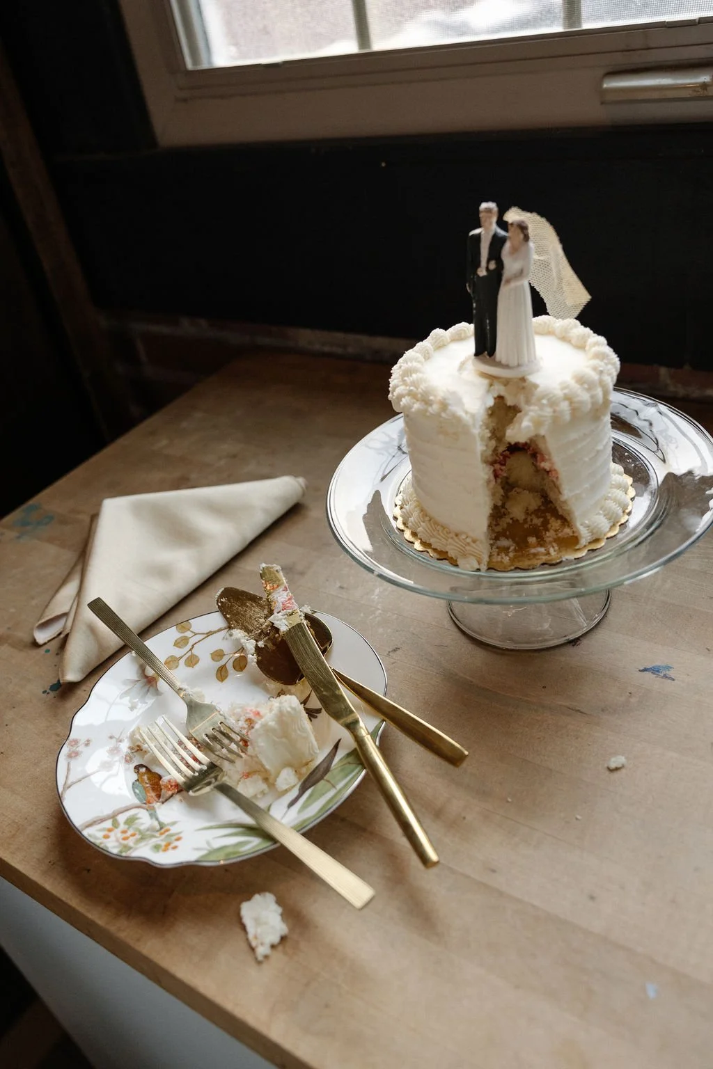 Partially eaten wedding cake with a bride and groom topper, on a glass cake stand; a plate with cake crumbs and utensils, and a cloth napkin on a wooden table near a window.