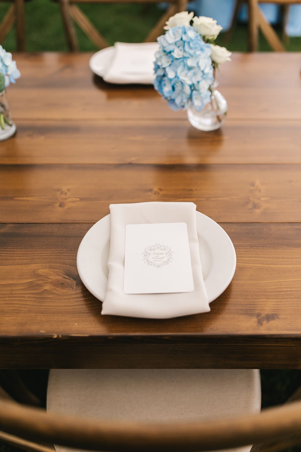 Elegant table setting with a white plate, a folded napkin, and a menu card, with a blue and white hydrangea floral centerpiece in the background.