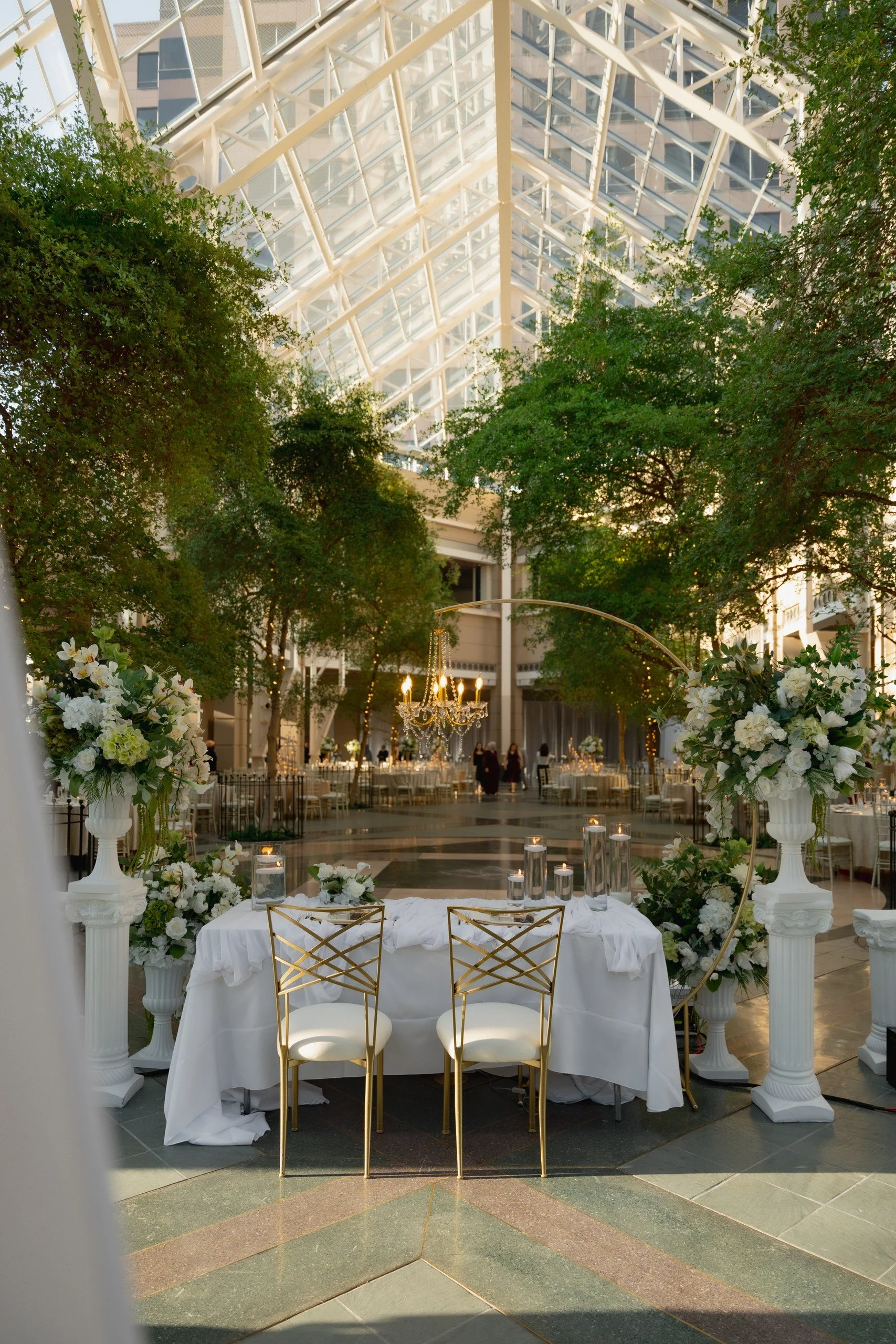 Elegant wedding reception setup inside a glass atrium with greenery, floral arrangements, and a white table with gold chairs.