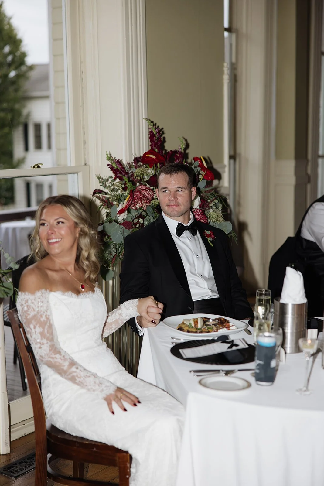 Bride and groom sitting at a wedding reception table, holding hands, with floral decorations in the background.