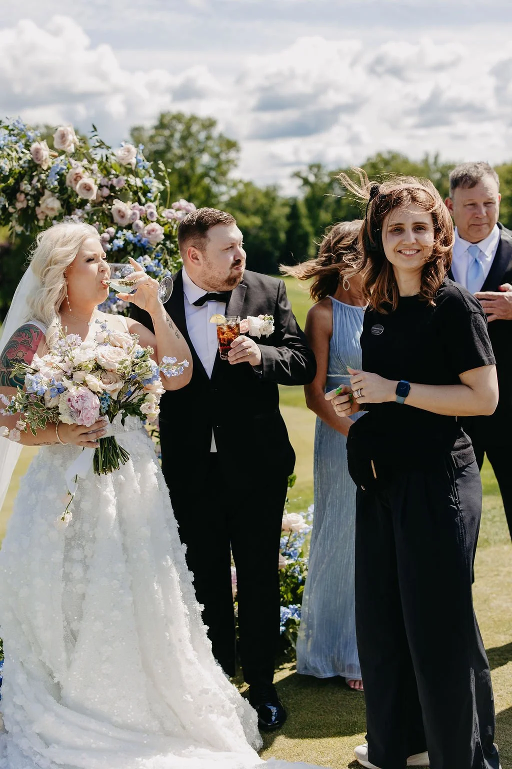 People at an outdoor wedding reception, with a bride holding a flower bouquet, drinking champagne, and guests in formal attire on a sunny day with a floral decoration in the background.
