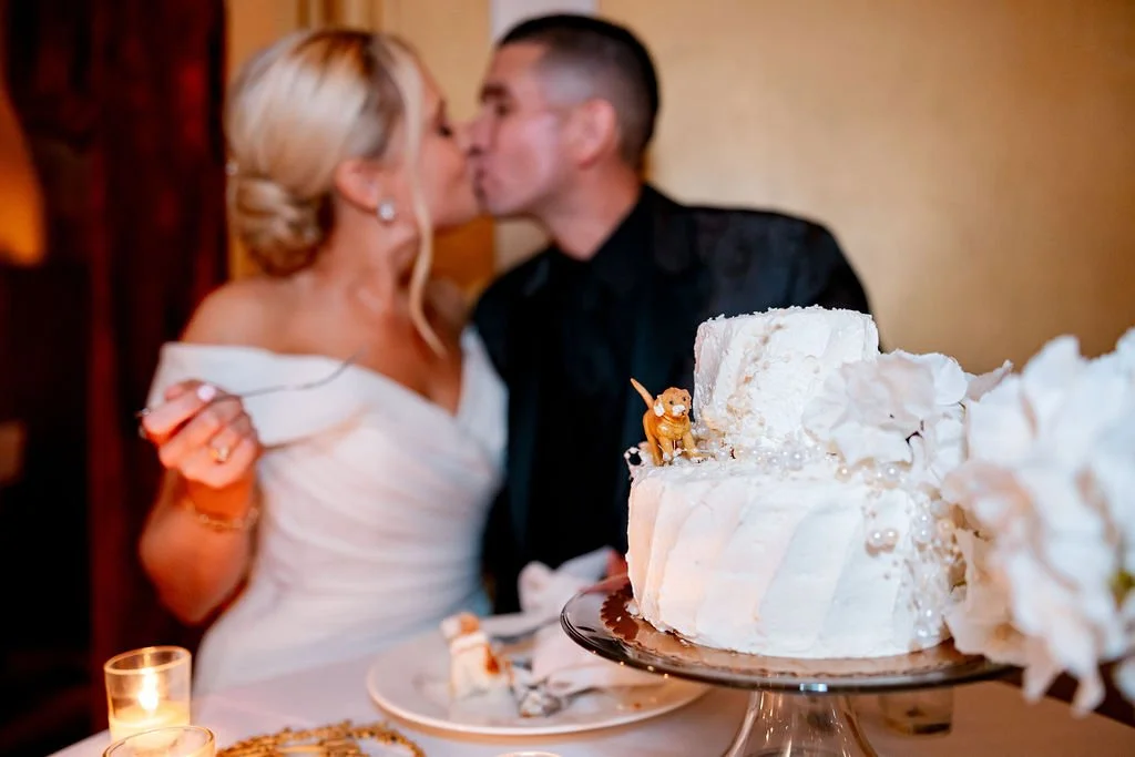 A bride and groom kissing during their wedding celebration, with a wedding cake in the foreground decorated with an animal figurine, flowers, and pearls.