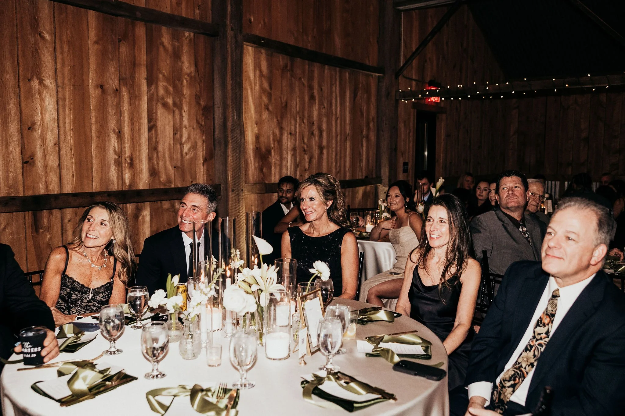 Guests in formal attire seated at a decorated banquet table in a rustic wooden venue, enjoying a celebratory event.