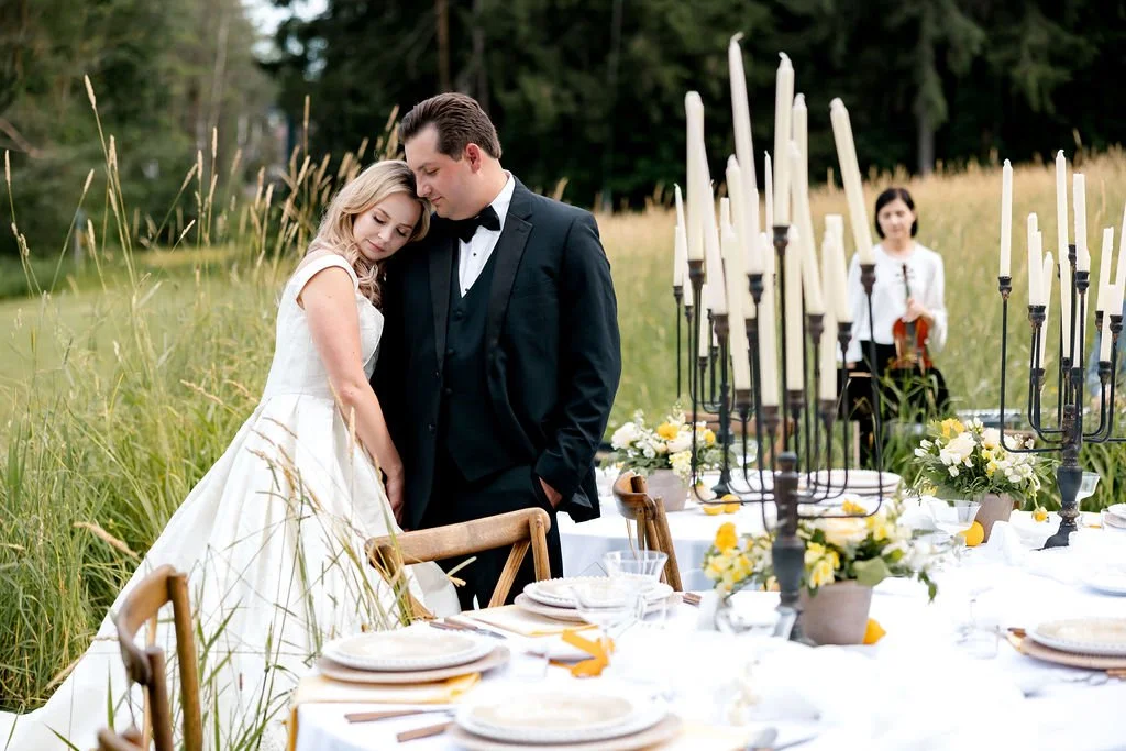 A bride and groom embracing outdoors at a wedding reception table in a field with tall grass and trees in the background, with a violinist playing in the distance.