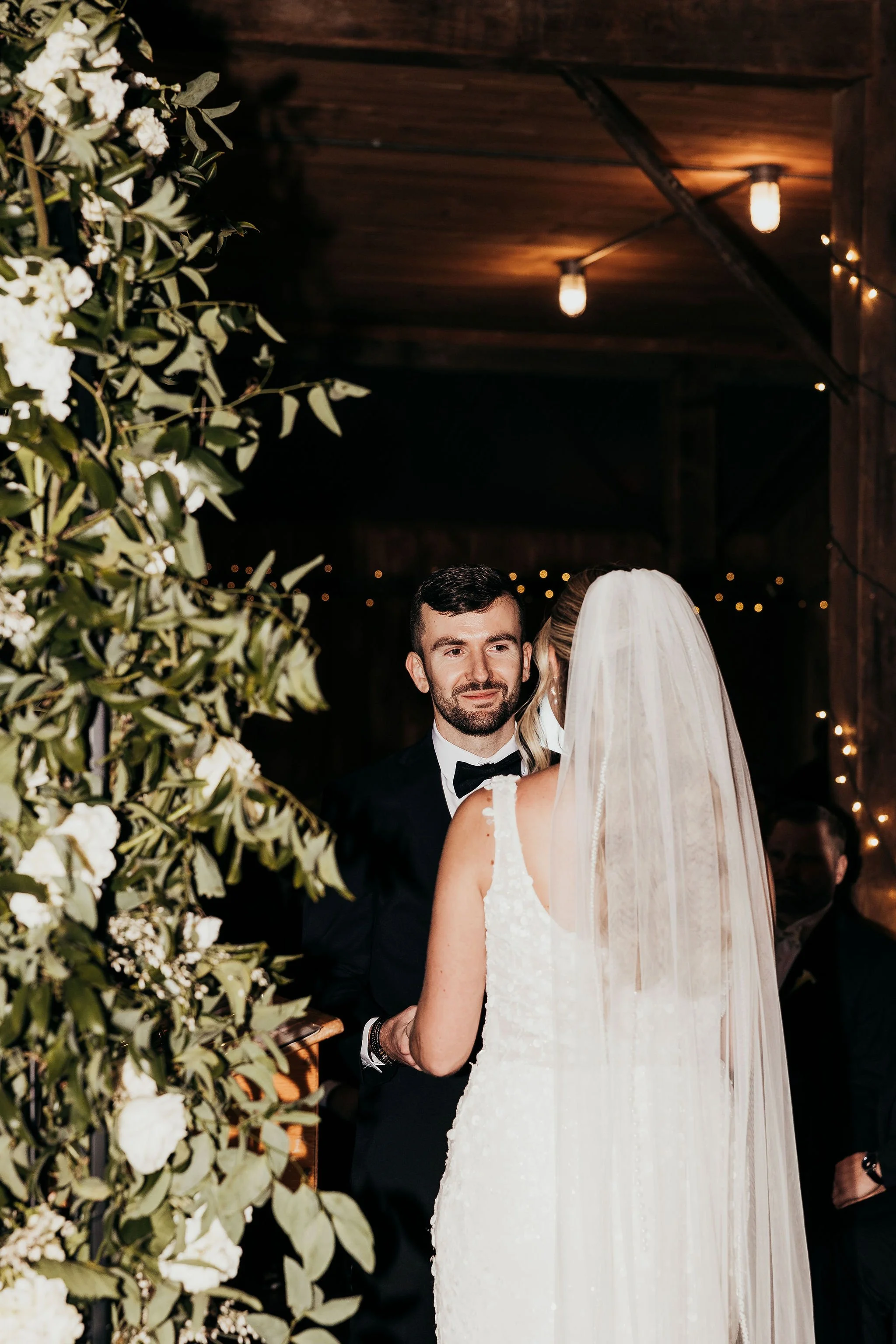A bride and groom exchanging vows at their wedding ceremony, with the bride in a white dress and veil, and the groom in a black tuxedo, surrounded by flowers and warm lighting.