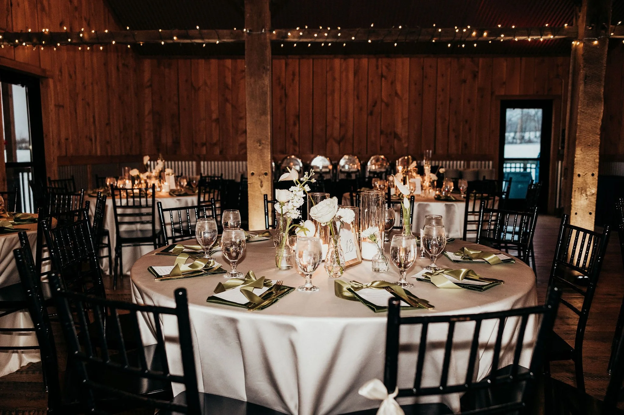 Round banquet table set for a formal event with white tablecloths, floral centerpieces, water glasses, napkins, and gold-colored bows, in a rustic wooden venue with string lights.